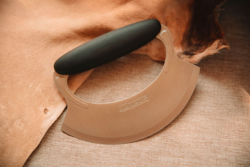 Mezzaluna dry scraper with curved stainless steel blade and black rubber handle on tanned hide and linen background