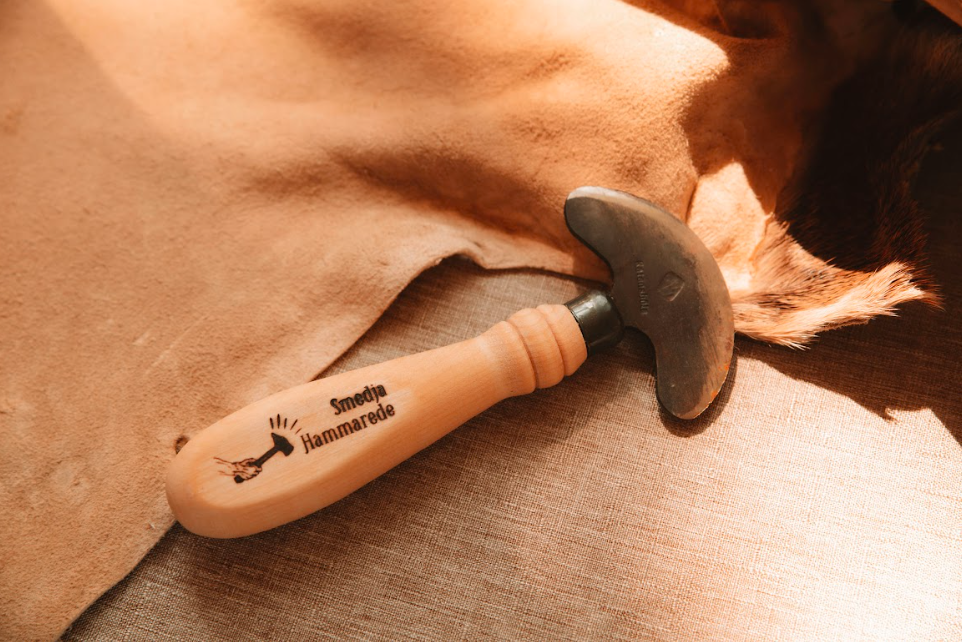 Hand-forged hide tanning scraper with rounded metal blade and lathed wooden handle resting on tanned hide and linen