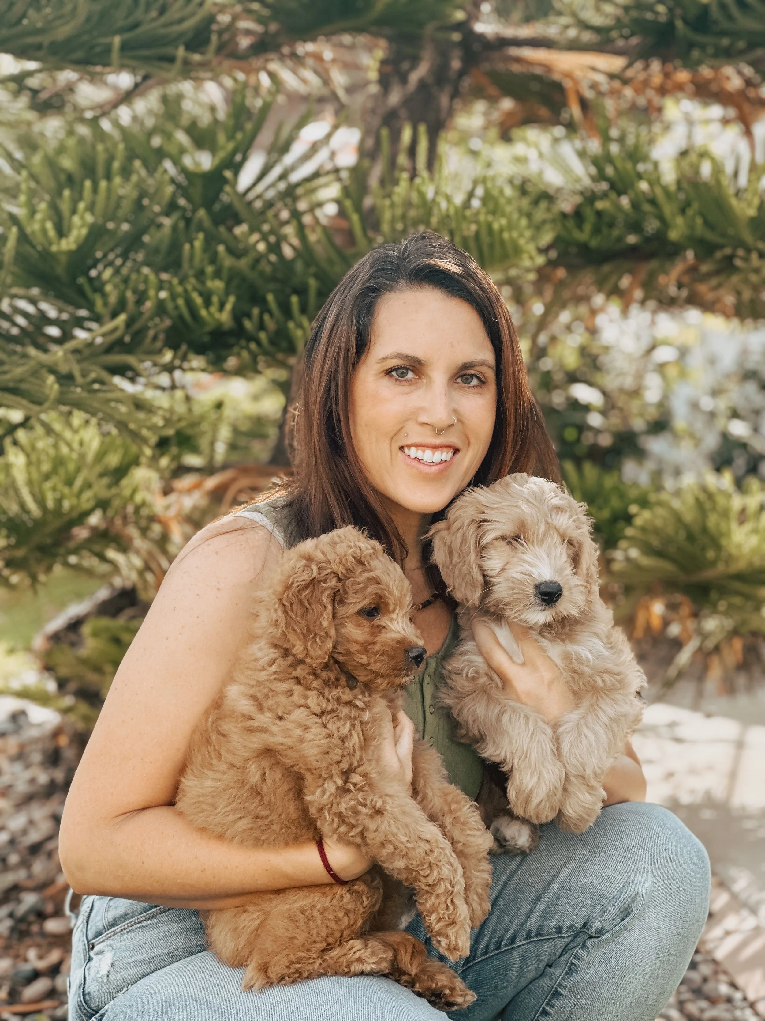 A woman with brown hair and a nose piercing holding two puppies outdoors with lush green plants in the background.