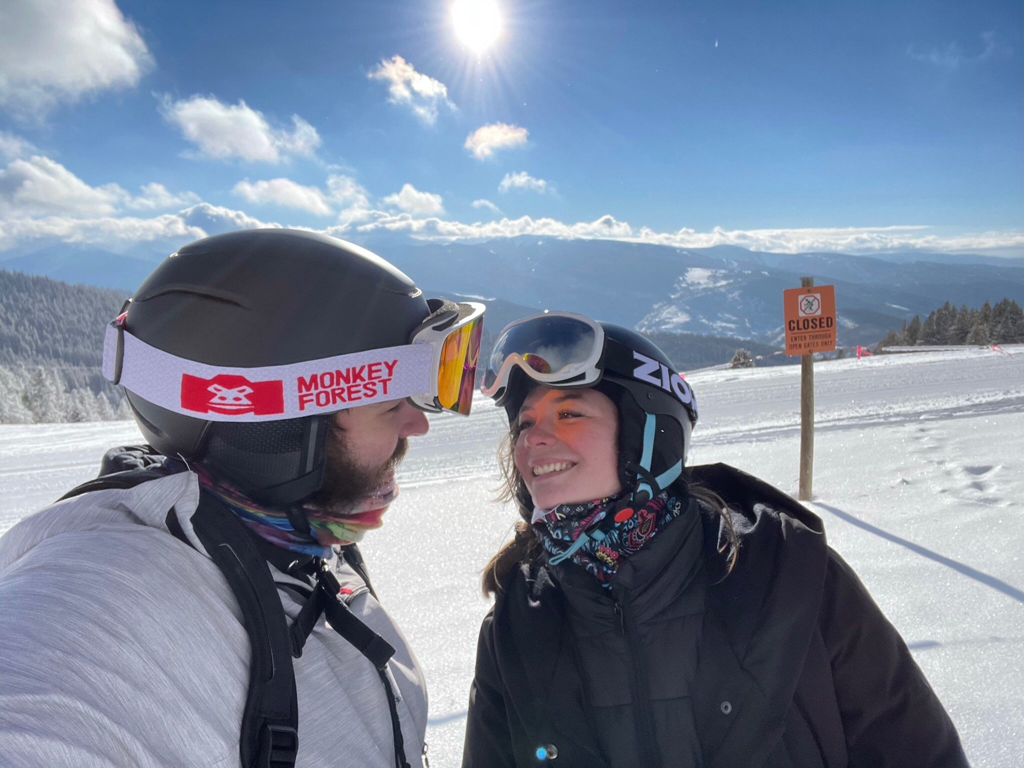 Two friends in winter gear smiling at each other on a snowy mountain with ski helmets and goggles, near a 'closed' sign, with a mountain landscape in the background.