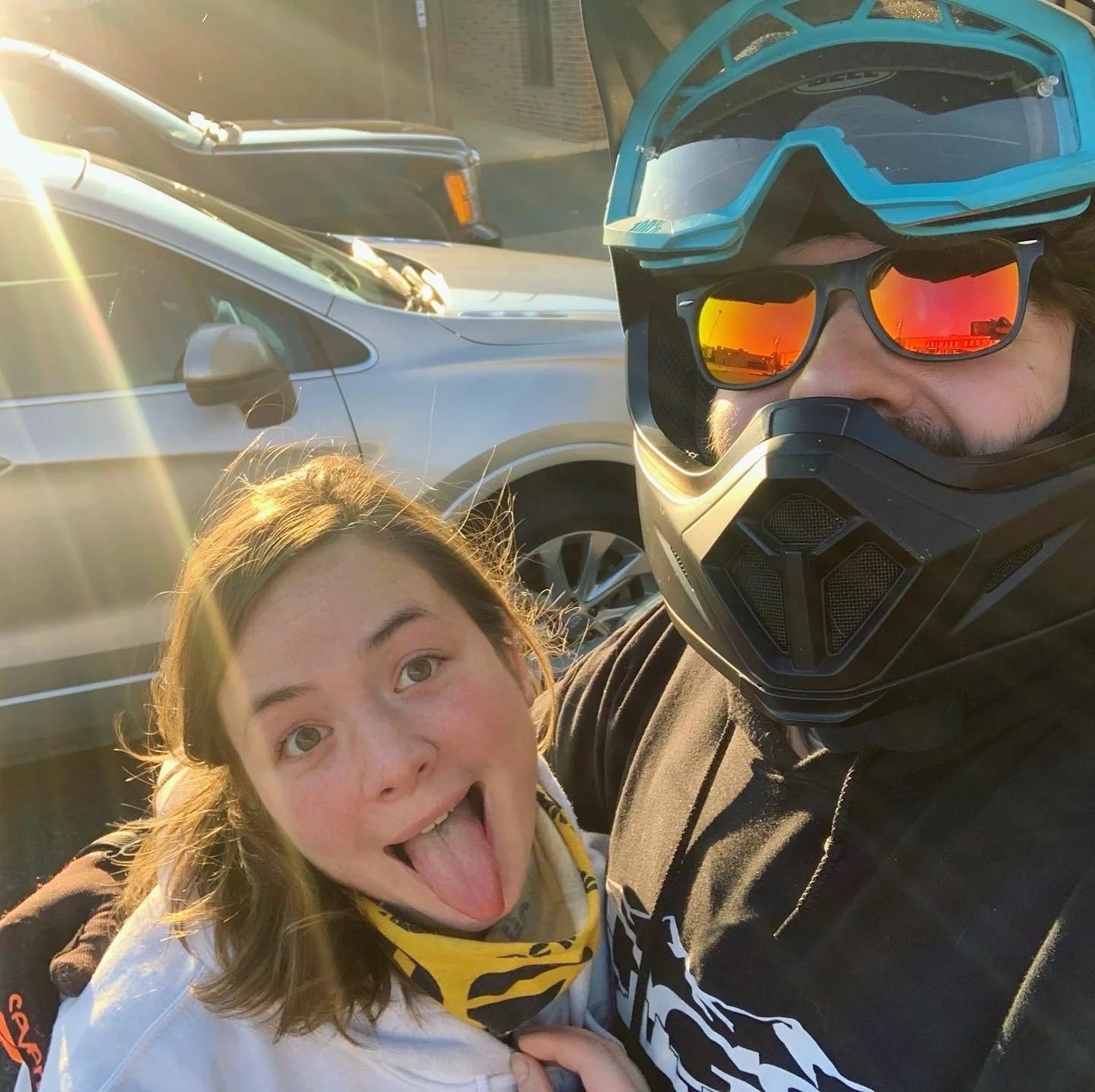 A woman sticking out her tongue and a man wearing a full-face motorcycle helmet and sunglasses taking a selfie together outside during sunset with parked cars in the background.