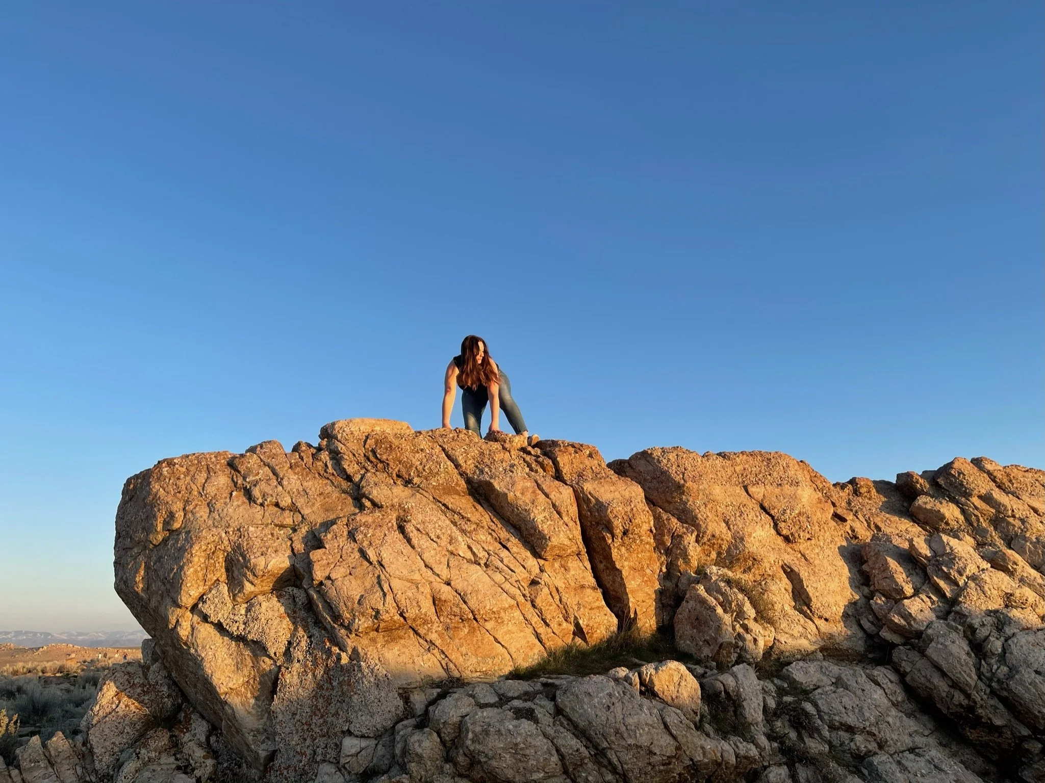 A woman climbing on a large rocky outcrop in a desert landscape during sunset with a clear blue sky.