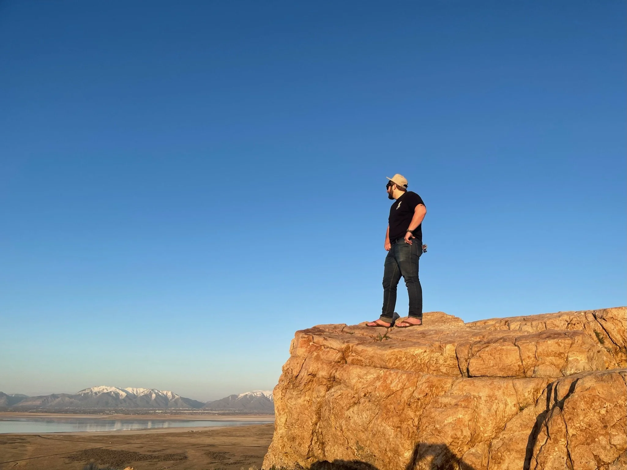 A man standing on a large rock formation overlooking a landscape with snow-capped mountains and a body of water during sunset.