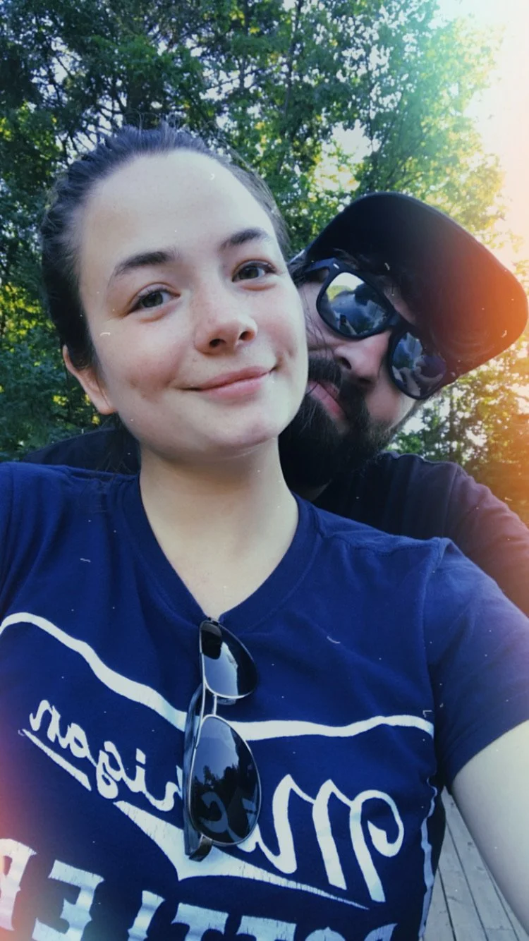 A young woman smiling at the camera, wearing a navy blue shirt with white text, sunglasses hanging from her shirt, with a man behind her wearing sunglasses and a cap. They are outdoors with trees and sunlight in the background.
