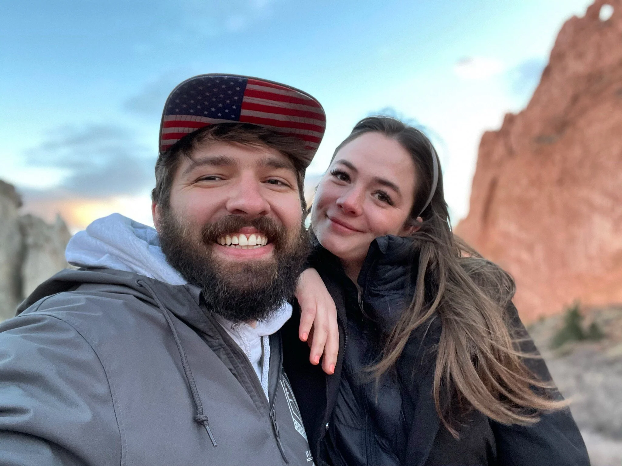 A smiling man with a beard and a woman with long hair taking a selfie outdoors with rock formations and a cloudy sky in the background.