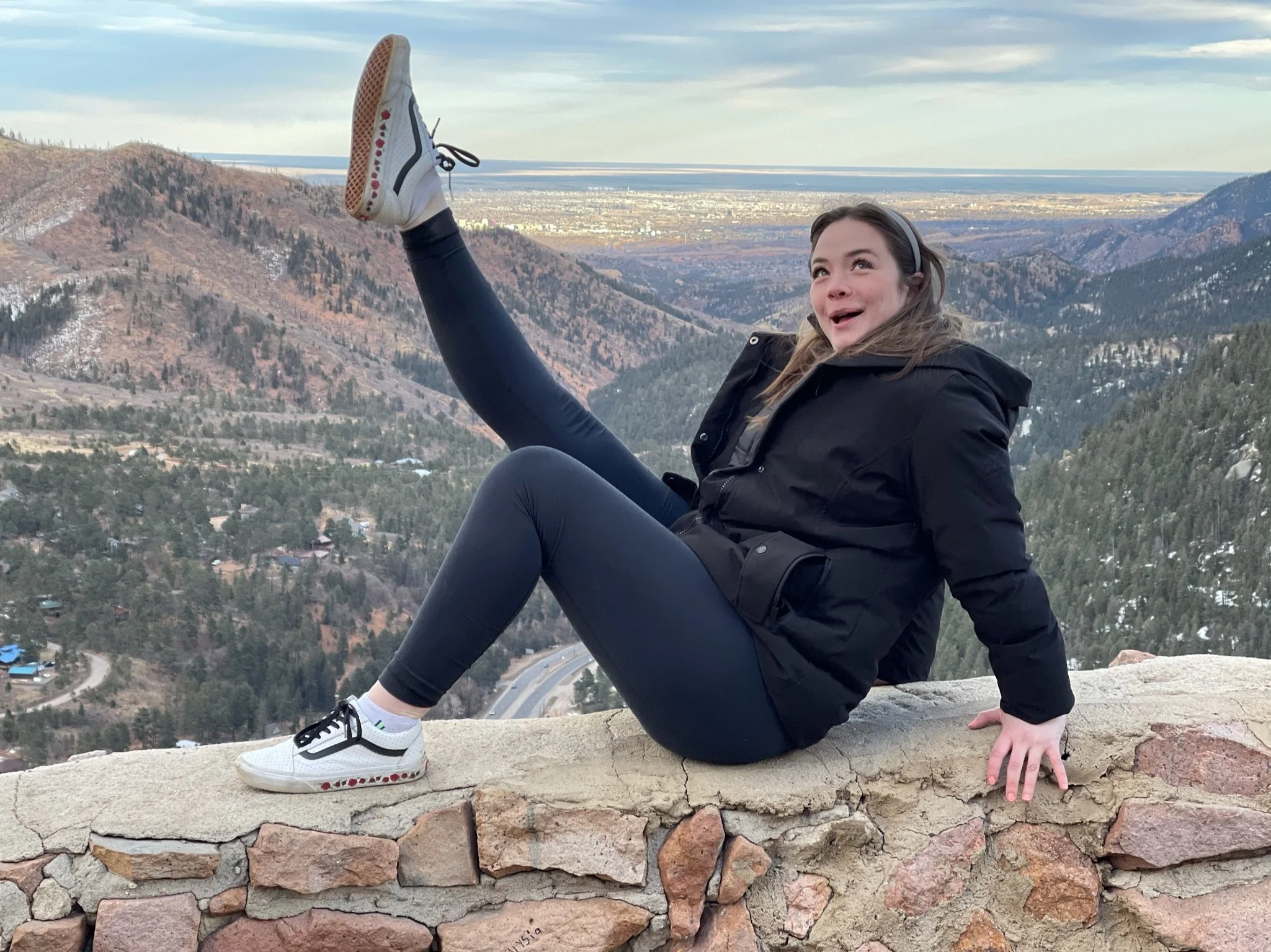 A young woman sitting on a stone wall with mountains and a valley in the background, smiling and raising one leg in the air.