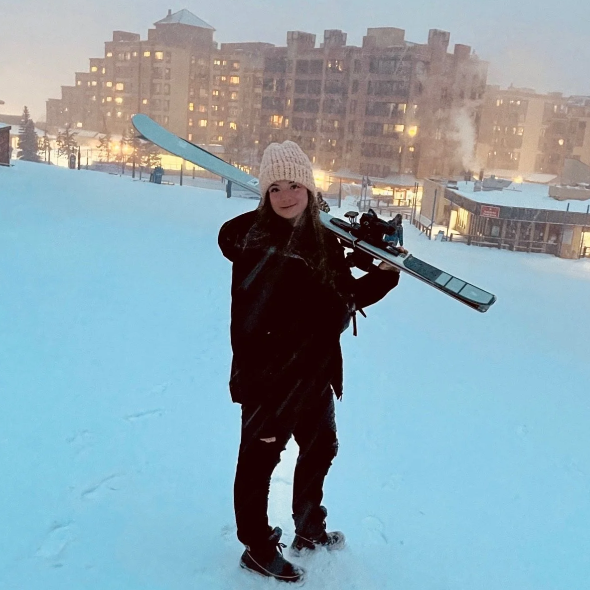 Woman in black winter jacket and white knit hat standing on snow with skis over her shoulder, in a snowy urban area at dusk, with buildings and lit windows in the background.