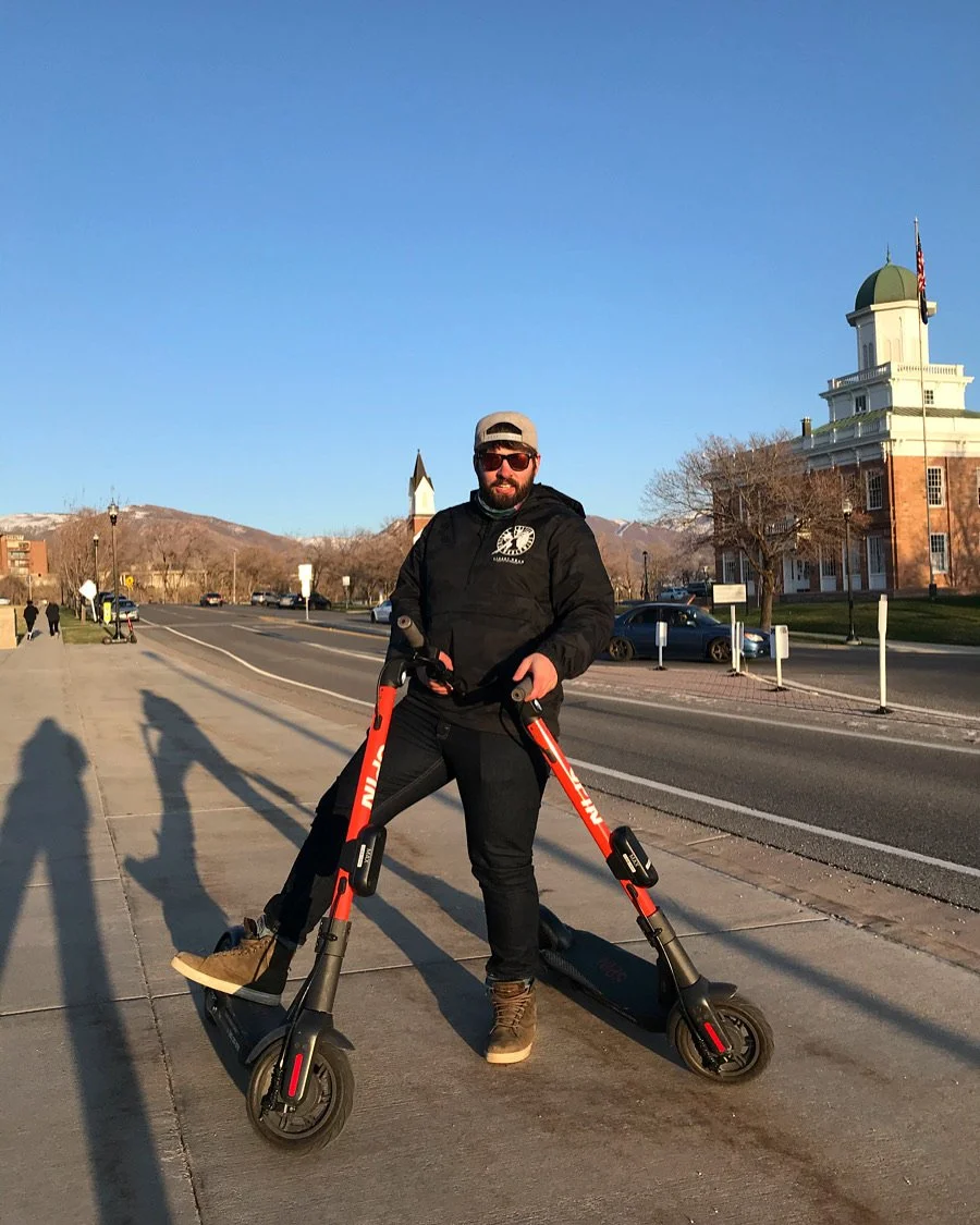 A man standing on an electric scooter on a sidewalk in a small town with buildings and mountains in the background.