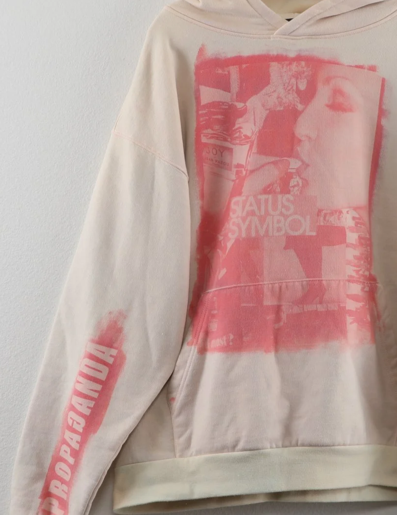 Cream-colored hoodie with pink graphic print of a woman's profile on the front and pink text on the sleeves. The hoodie hangs on a black hanger on a white wall.