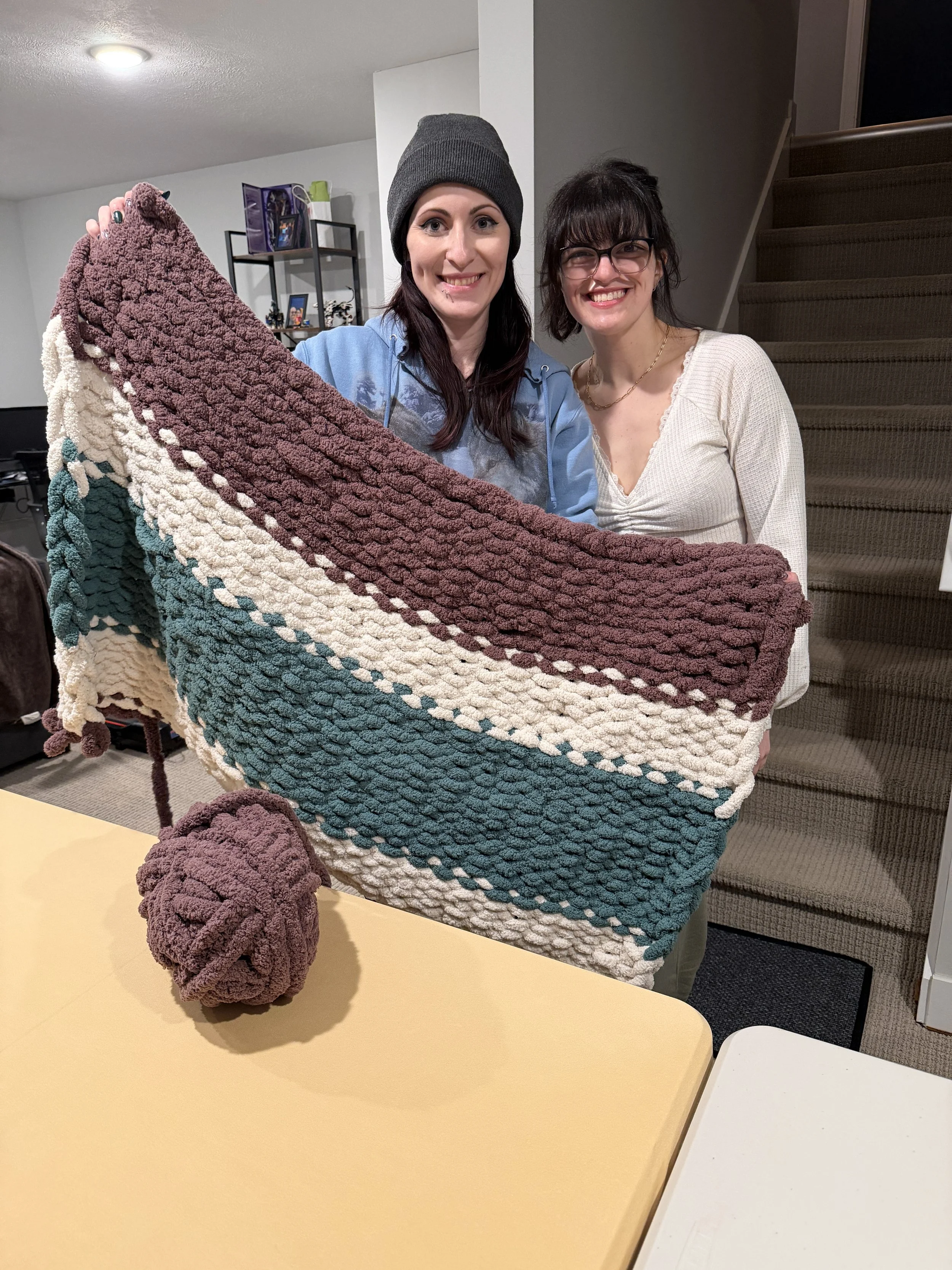Two white women holding a knit blanket wearing a white shirt and blue sweatshirt.