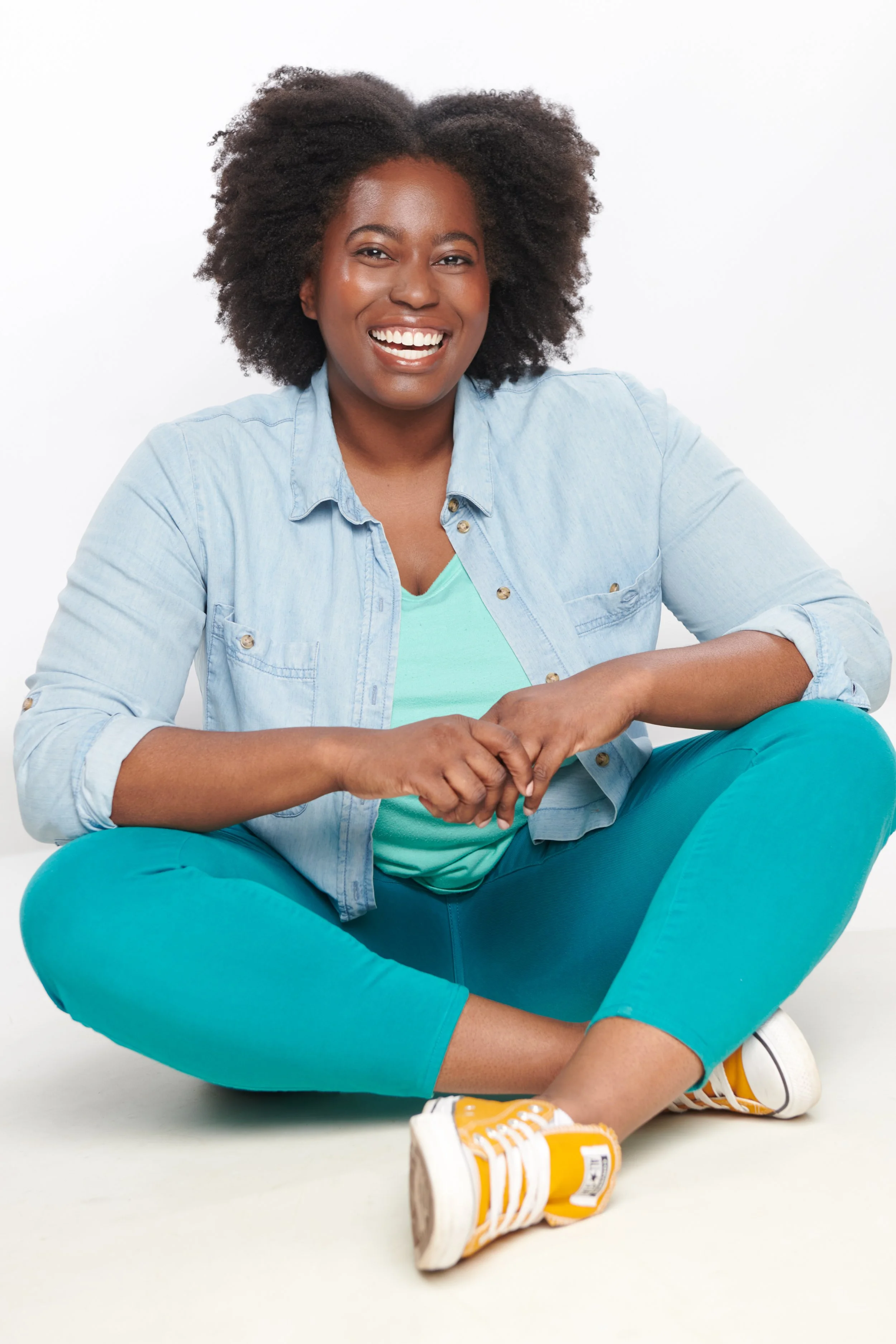 A smiling Black woman sitting cross-legged, wearing a denim shirt, turquoise pants, and yellow sneakers, against a white background.