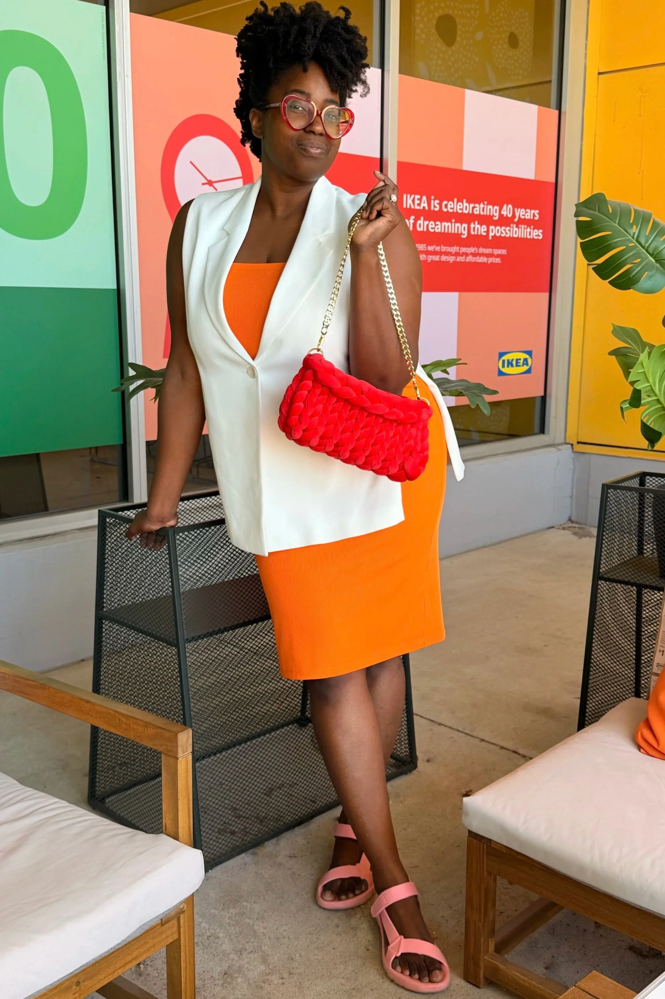 A woman with curly hair, wearing glasses and a white sleeveless blazer over an orange dress, posing inside an IKEA store. She has a red braided handbag and is wearing pink sandals. The background features IKEA promotional signs and indoor plants.