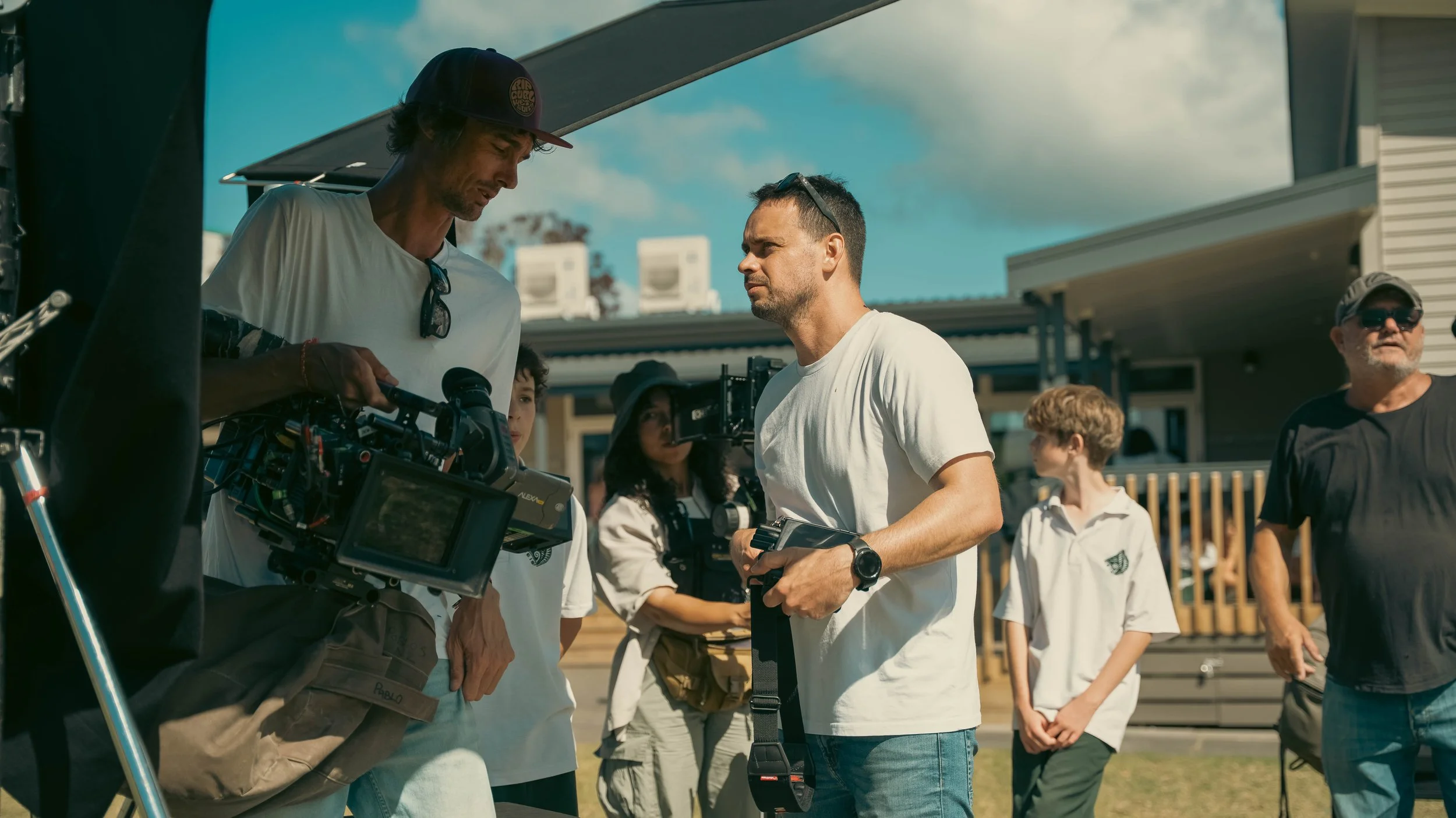 Filmmakers and actors on set filming outdoors, with crew members and equipment visible, at a sunny residential location.