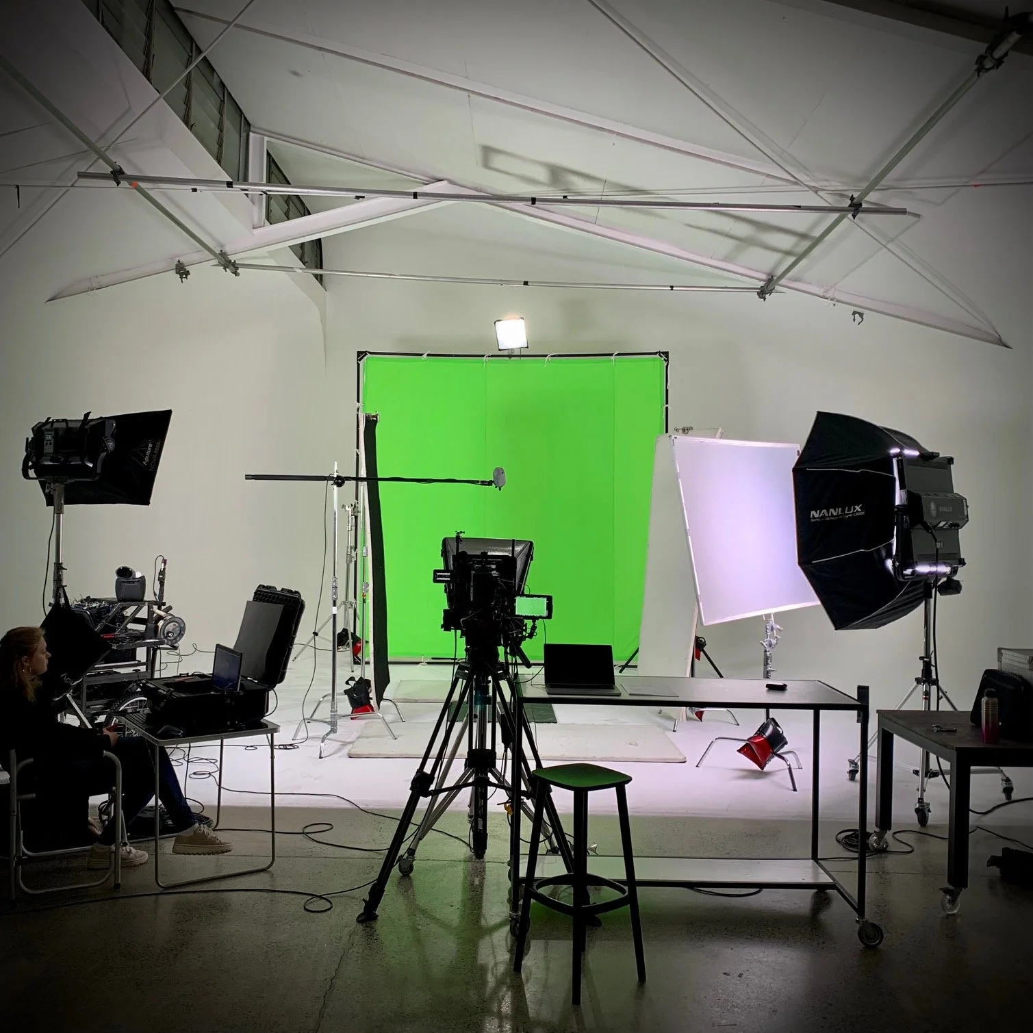 A professional photography or video studio with a green chroma key backdrop, lighting equipment, cameras, and other studio gear.