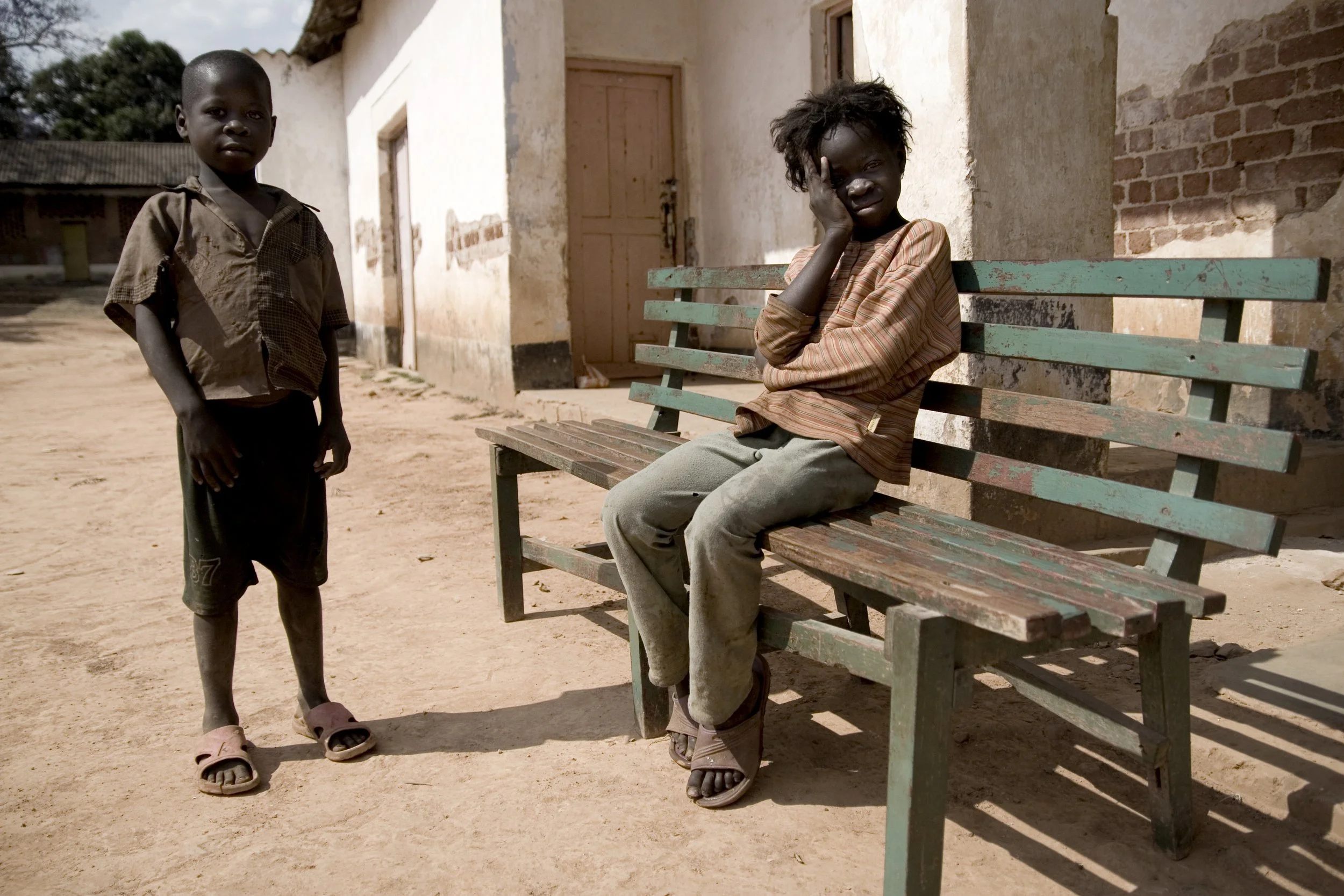 A young boy standing next to a girl sitting on a weathered park bench outside a building with weathered walls.