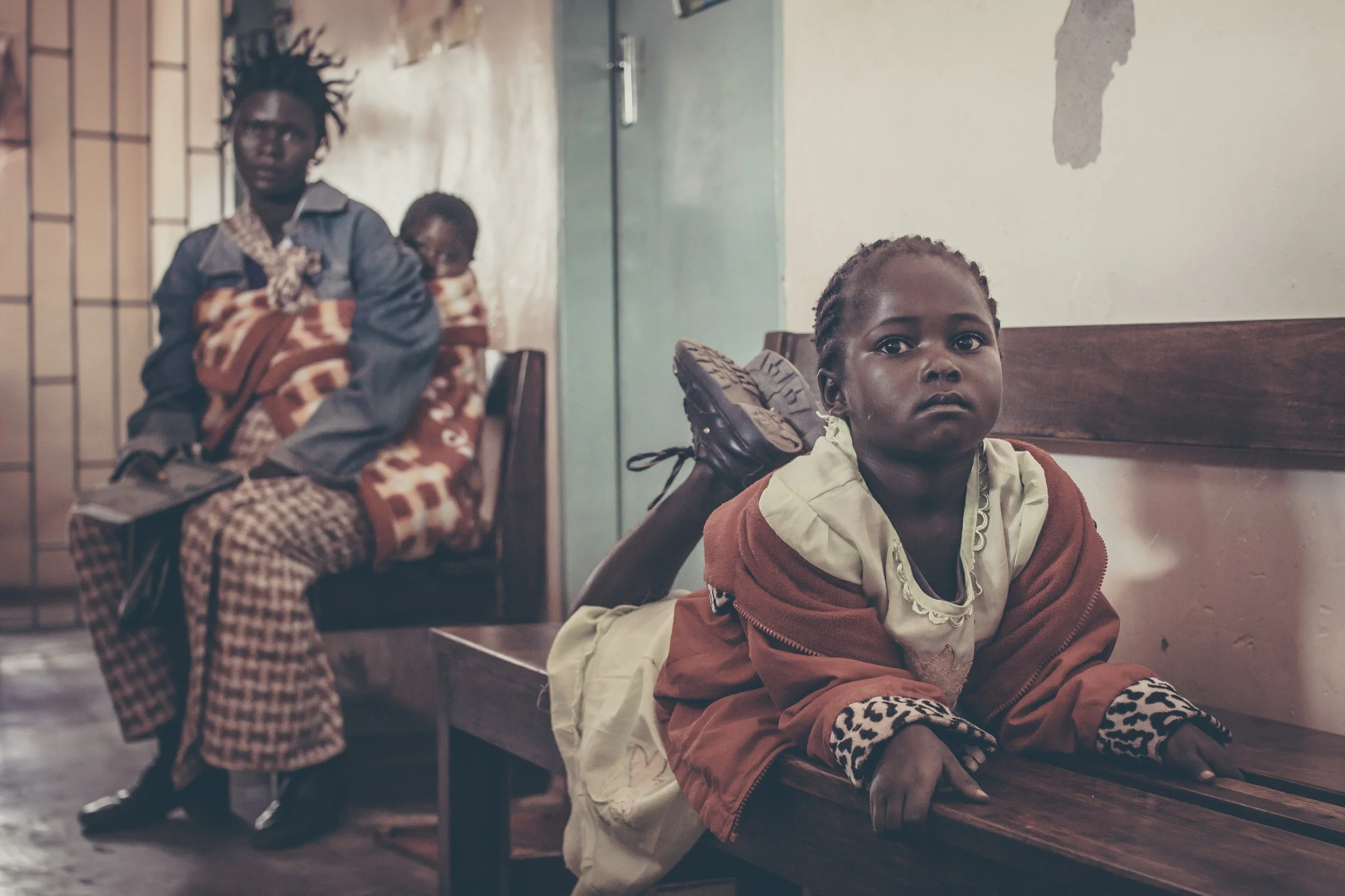 A young girl lying on her stomach on a wooden bench, looking at the camera. Behind her, two women are sitting on chairs, one holding a child wrapped in a blanket. The setting appears to be indoors, possibly in a clinic or waiting area.