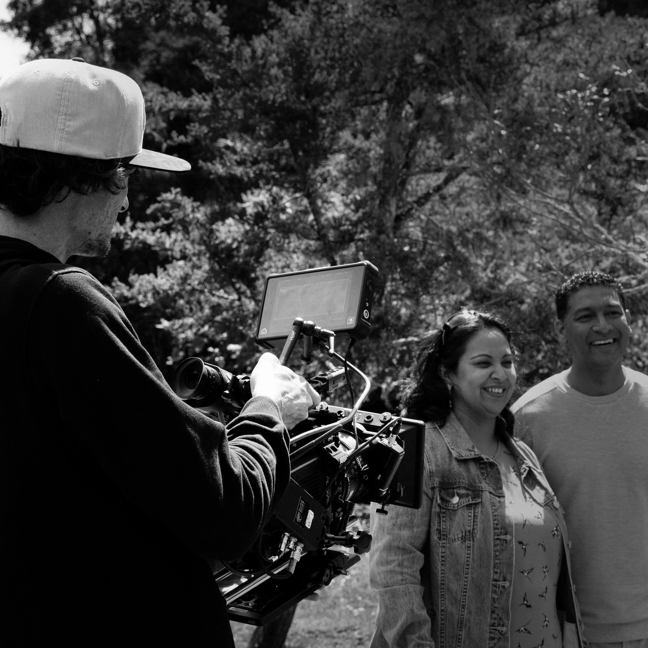 A person operating a film camera outdoors with two smiling people standing next to them, trees in the background.
