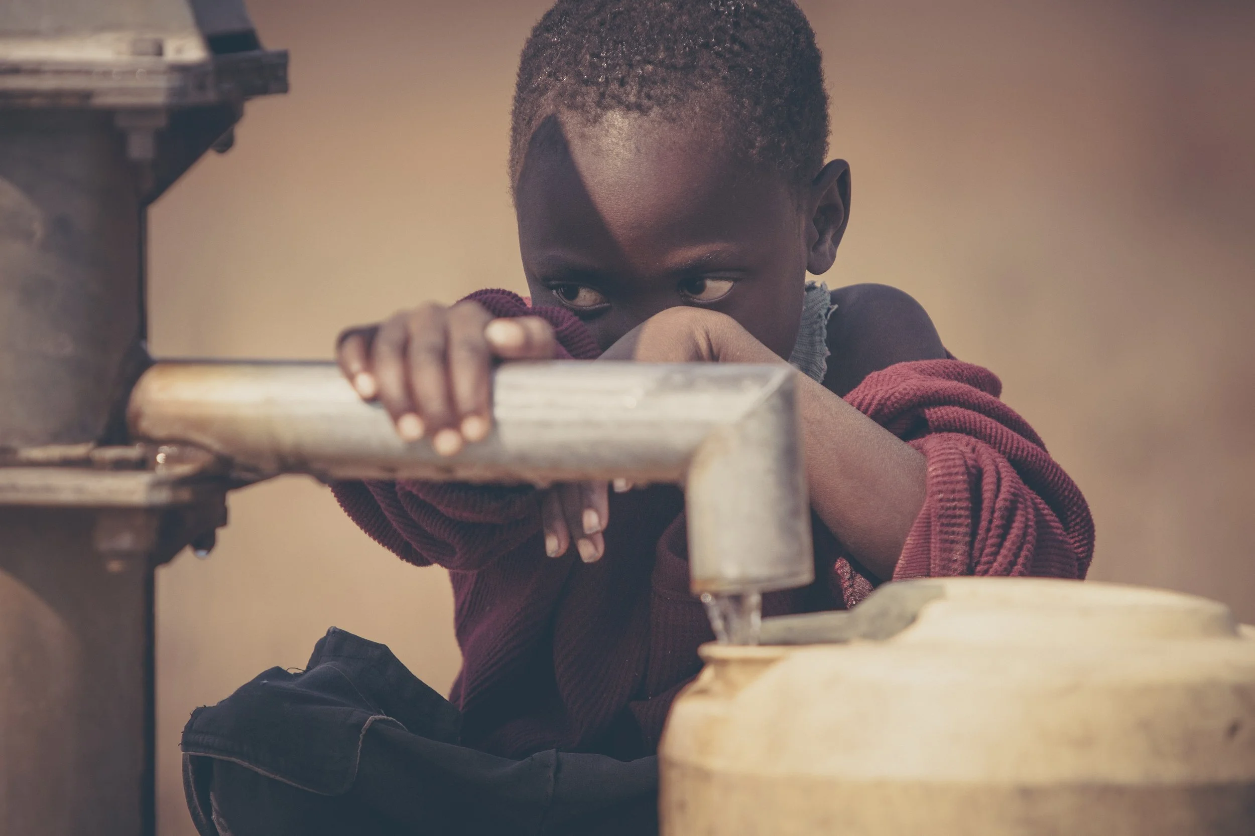 A young boy with short curly hair, wearing a red and black long-sleeved shirt, is leaning on a water fountain, drinking from it with his eyes looking to the side.