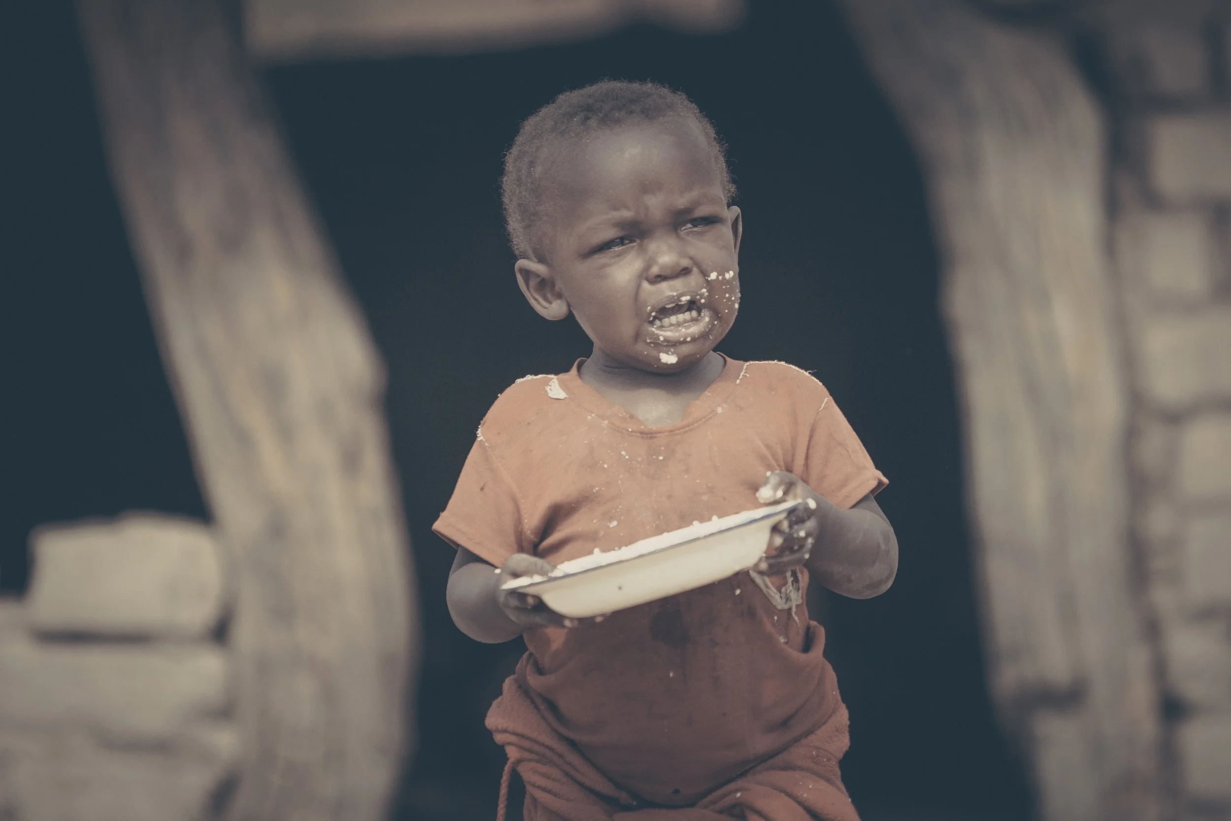 A young child holding a bowl with some food in an outdoor setting, with a dark background and tree trunks visible on either side.