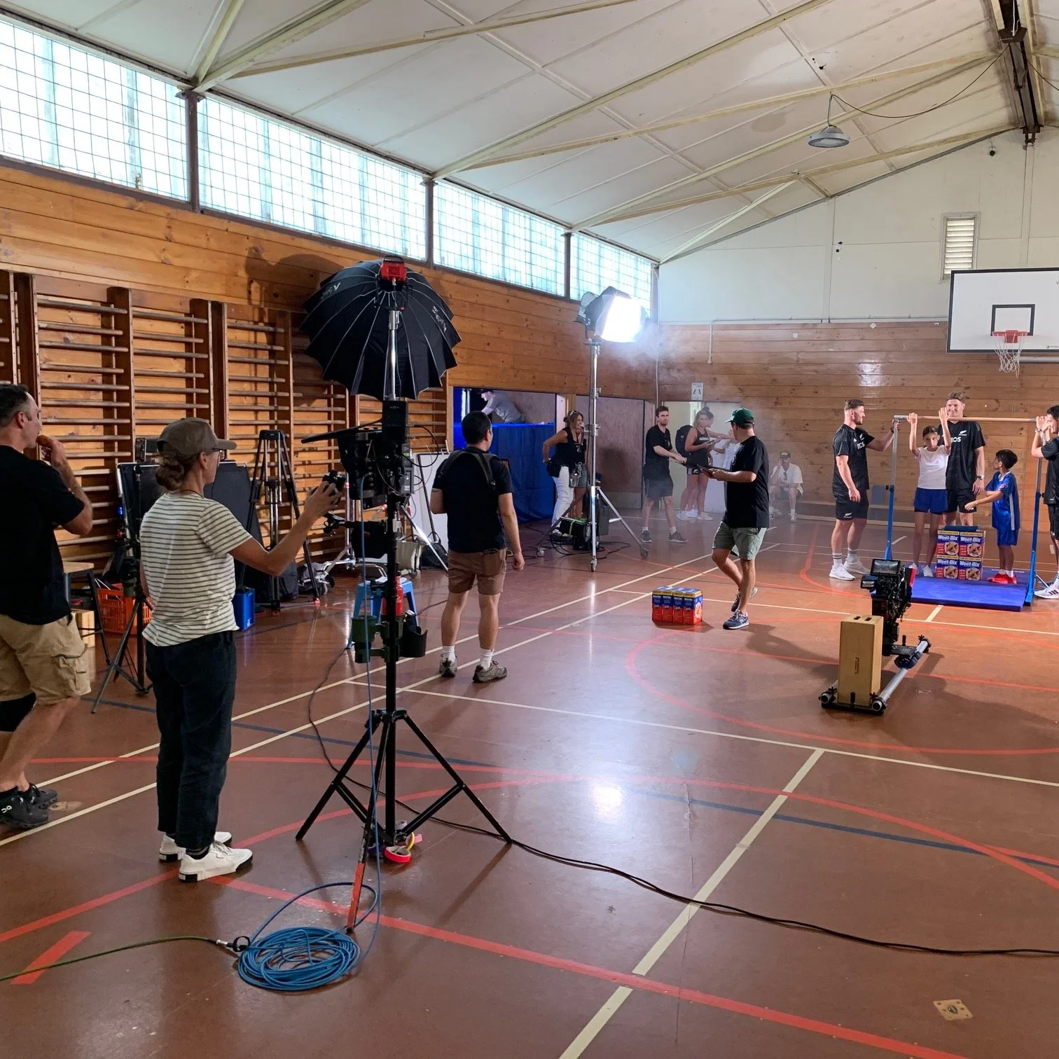 Behind the scenes of a sports video shoot in a gymnasium with filming equipment, crew members, and children practicing gymnastics.