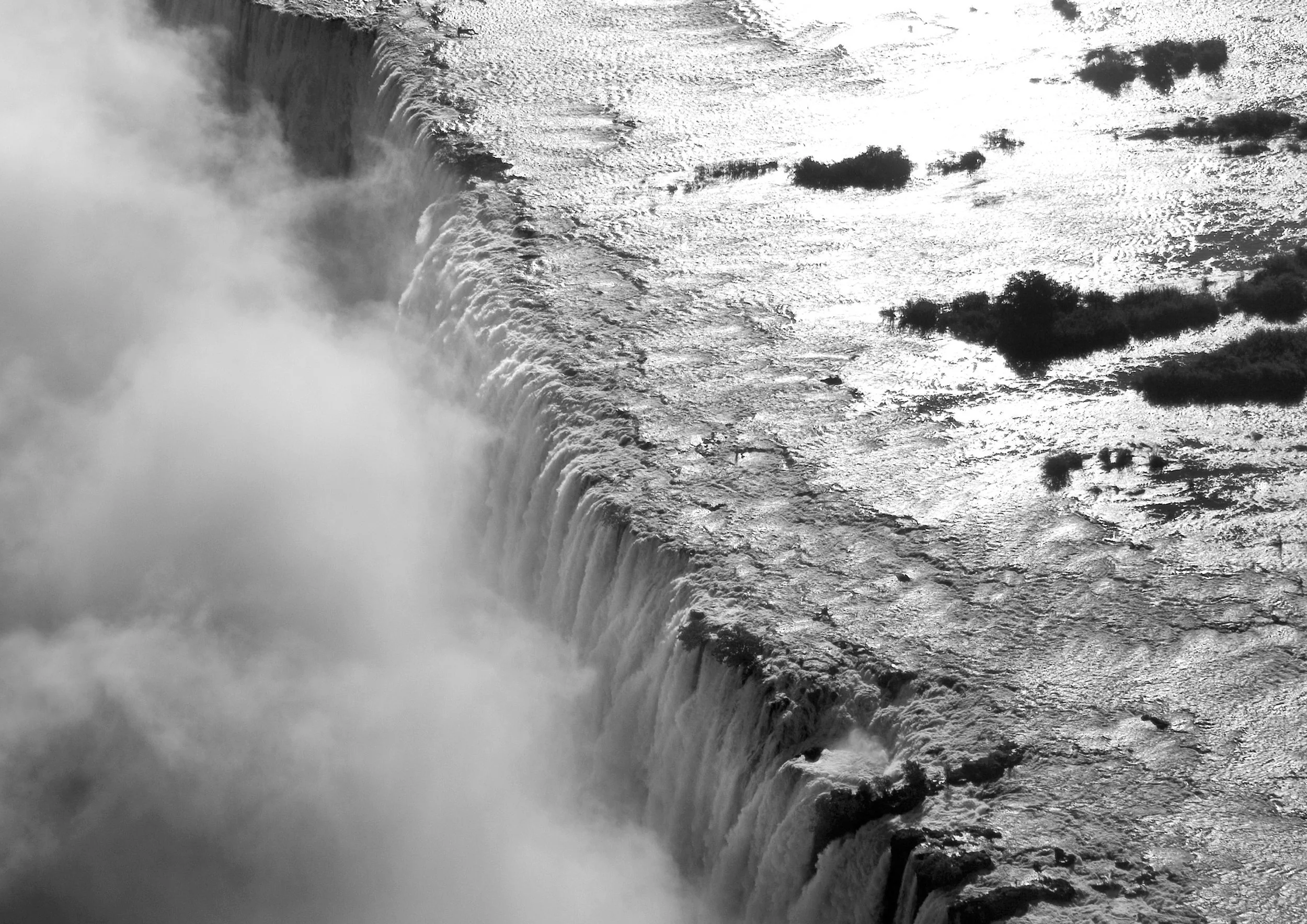 Black and white photograph of Niagara Falls showing cascading water and mist rising from the falls with rocks in the water.