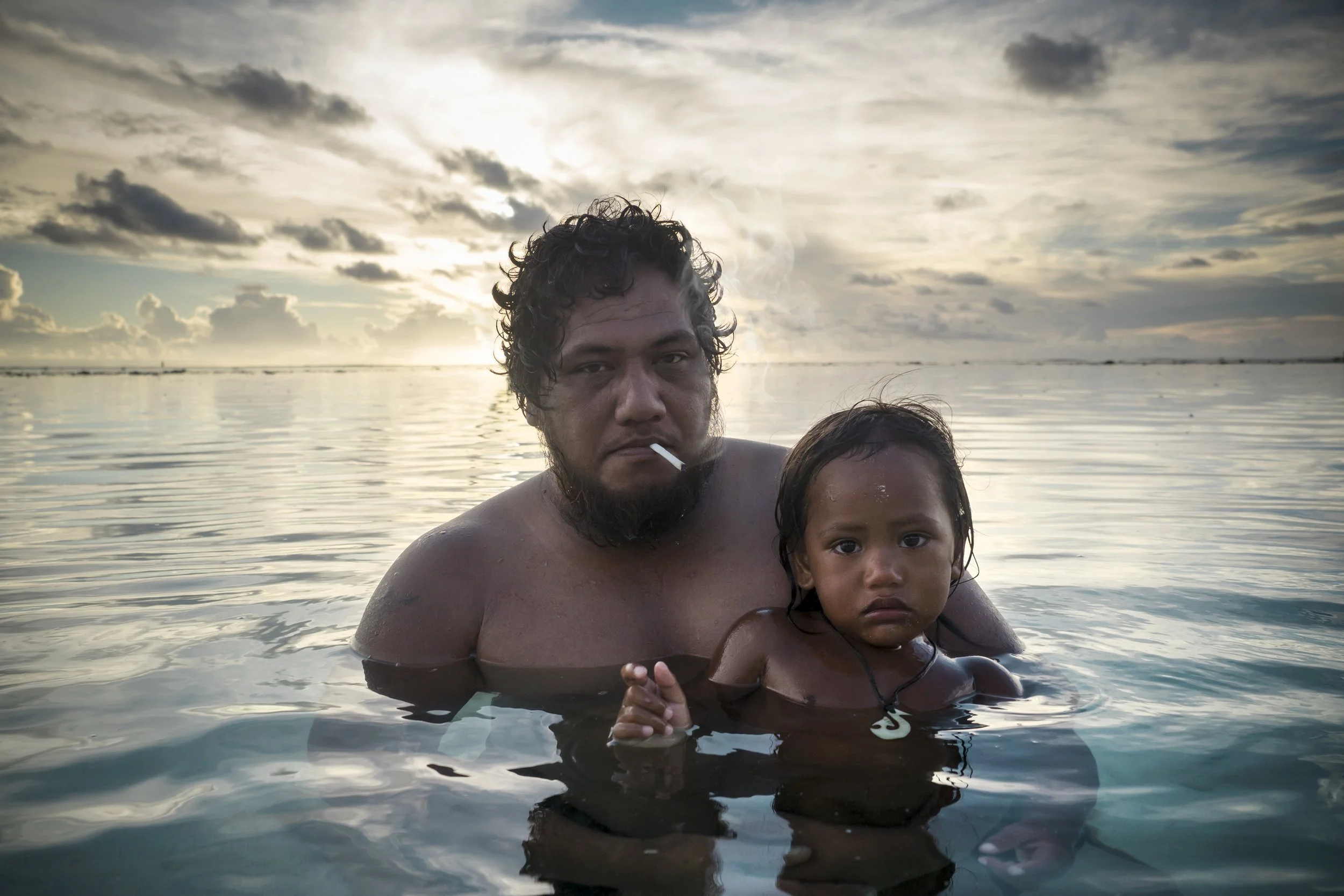 A man and a young girl are swimming in the ocean during sunset, with cloudy skies in the background.