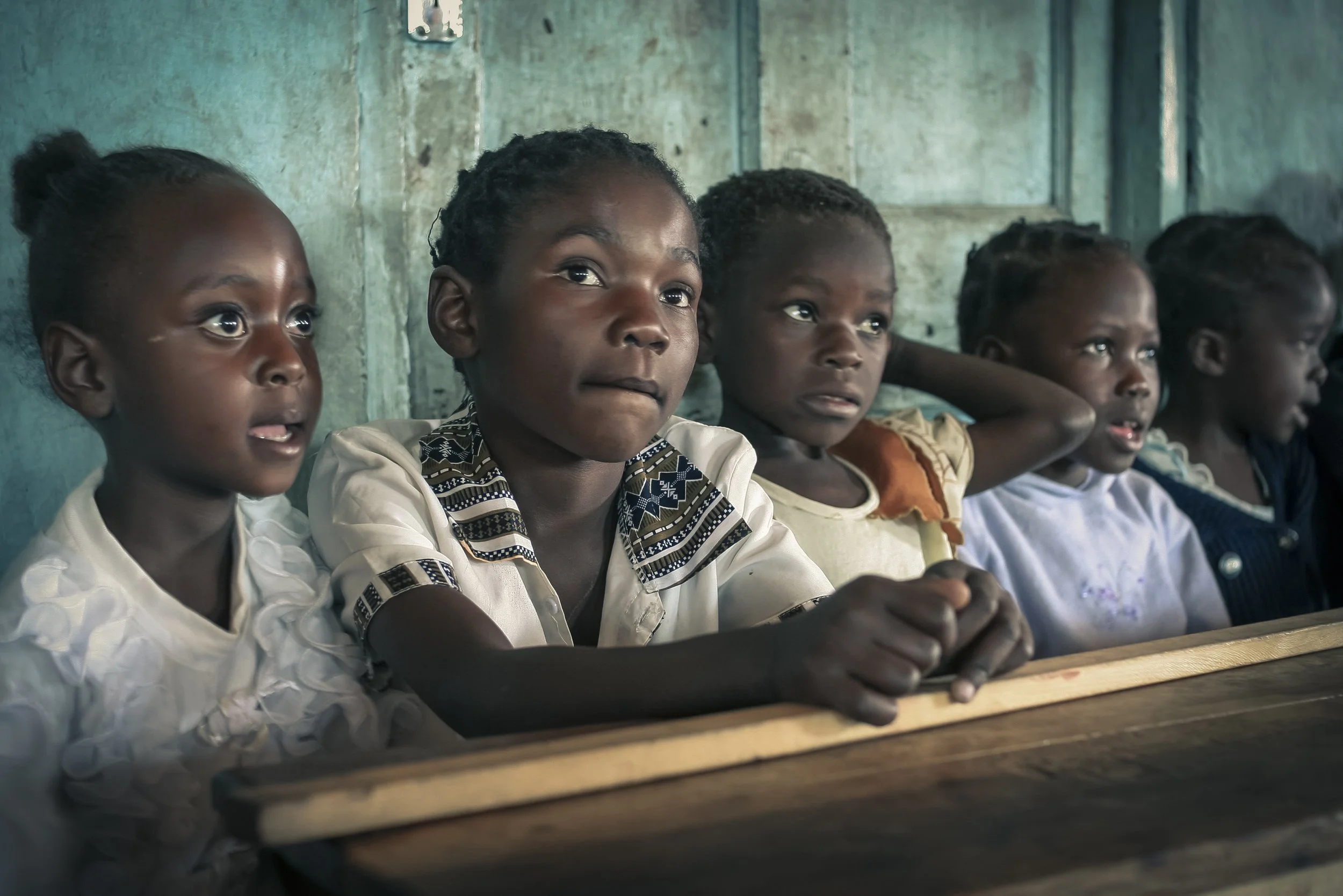 Five young African girls sitting at a wooden desk inside a classroom, attentively listening.