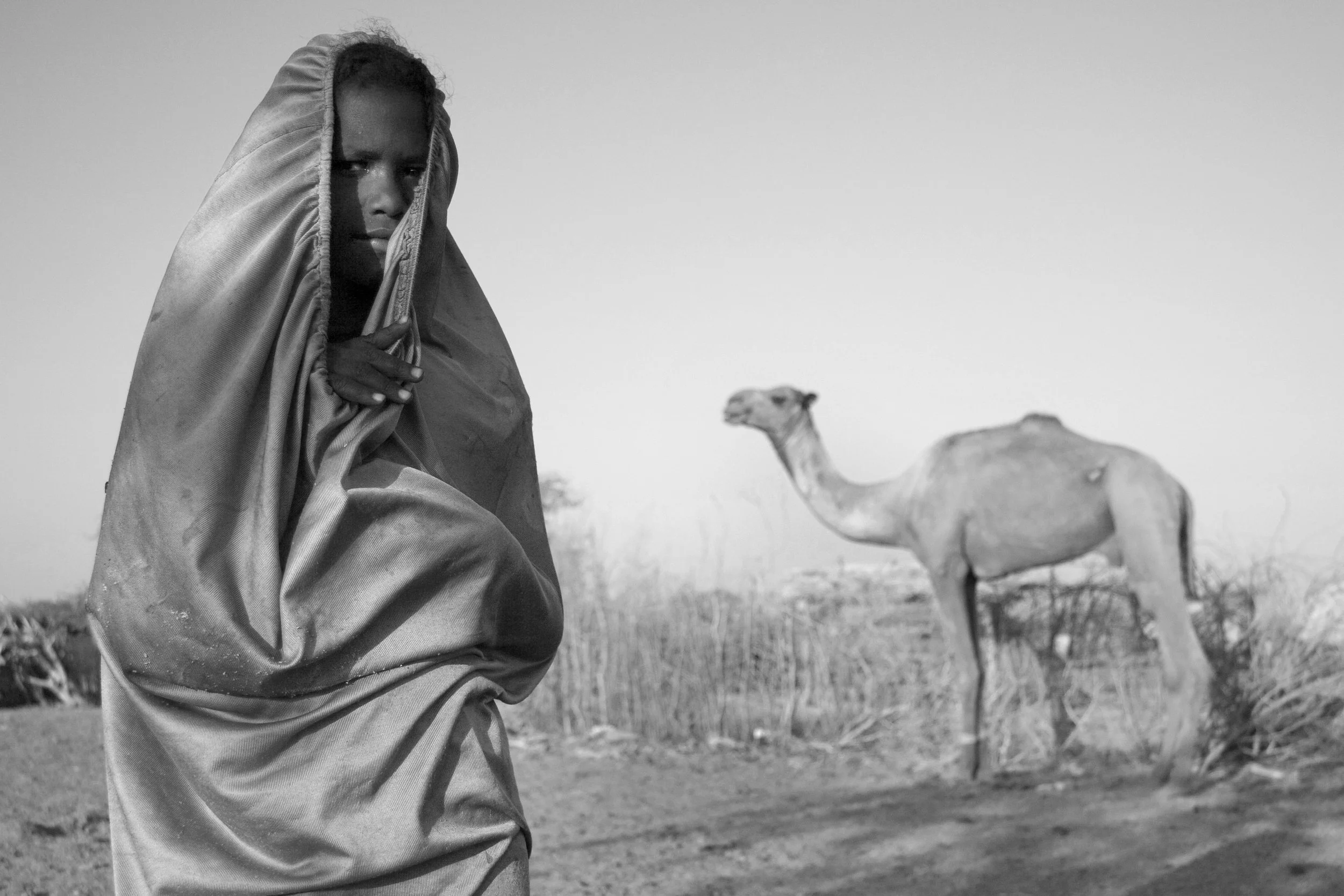 A person with dark skin and a serious expression, wrapped in a hooded cloth, stands in front of a camel in a desert landscape.