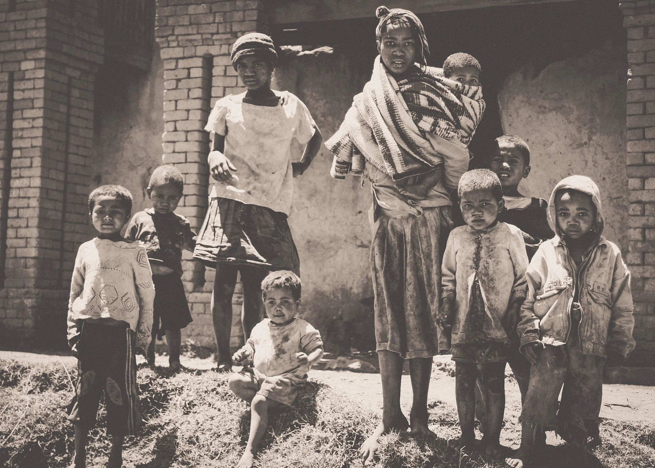 A black-and-white photo of a woman with children outside a brick building. The woman is carrying a young child on her back, wrapped in a blanket, and there are several children standing or sitting around her.