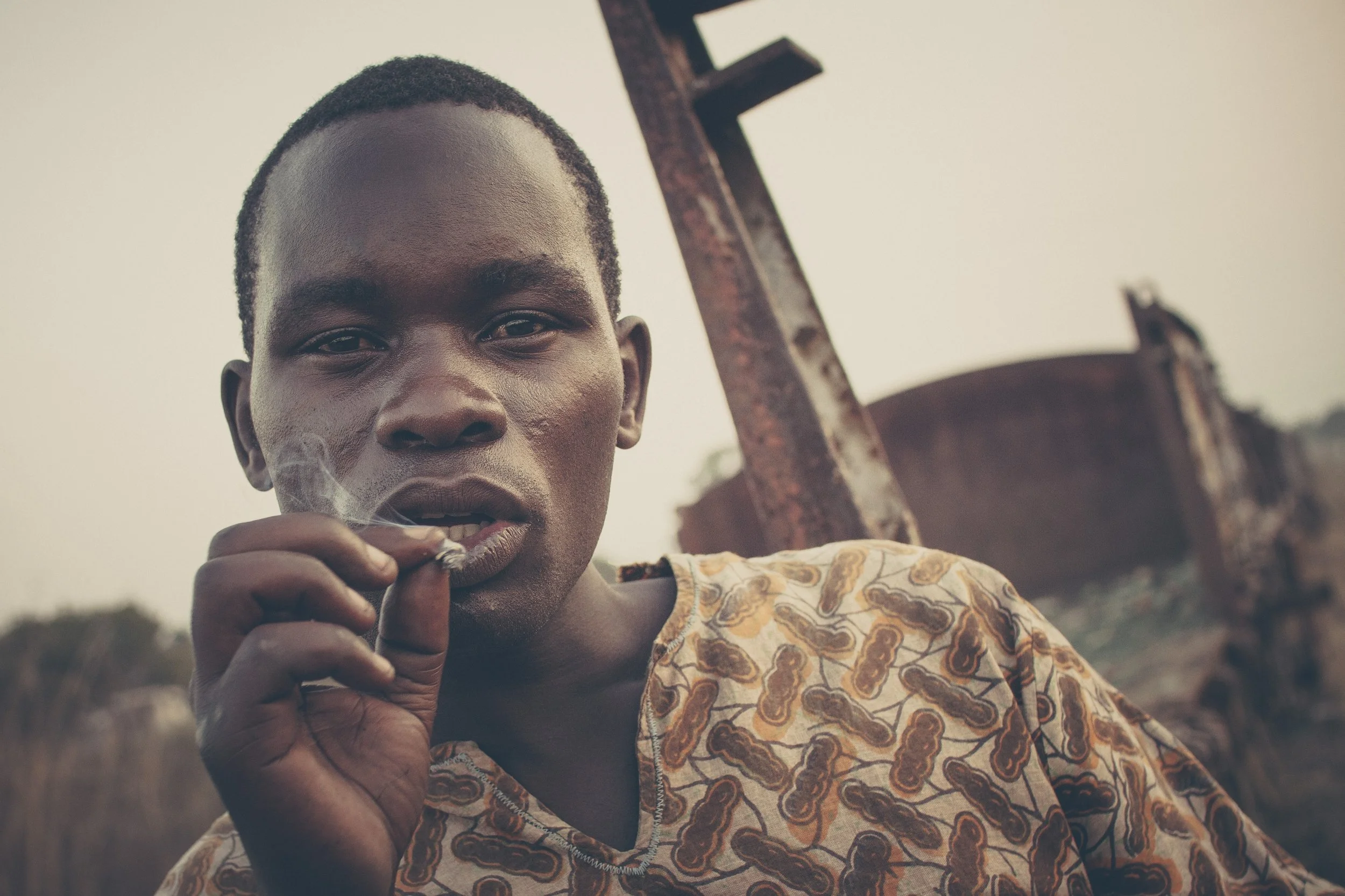 Young man with short hair smoking a cigarette outdoors, wearing a patterned shirt, with a rusty object in the background.
