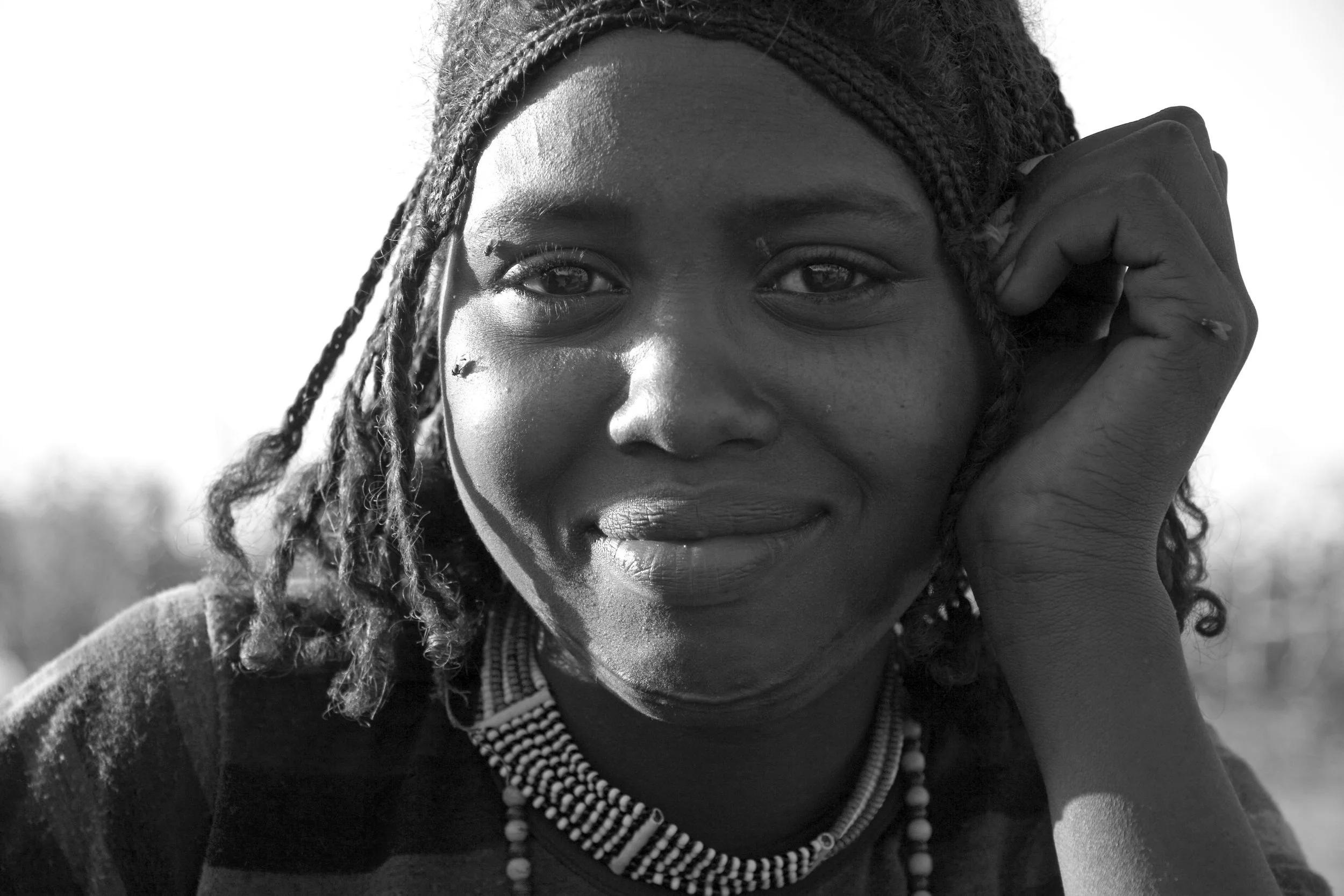 Close-up black and white portrait of a woman with braided hair wearing beaded necklaces and smiling softly, resting her head on her hand.