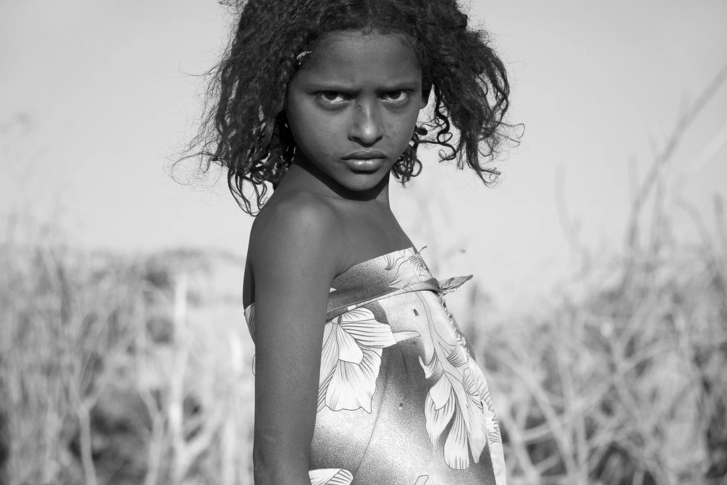 A young girl with curly hair, dressed in a floral-patterned dress, looks intensely at the camera in a black and white outdoor setting with dry grass in the background.