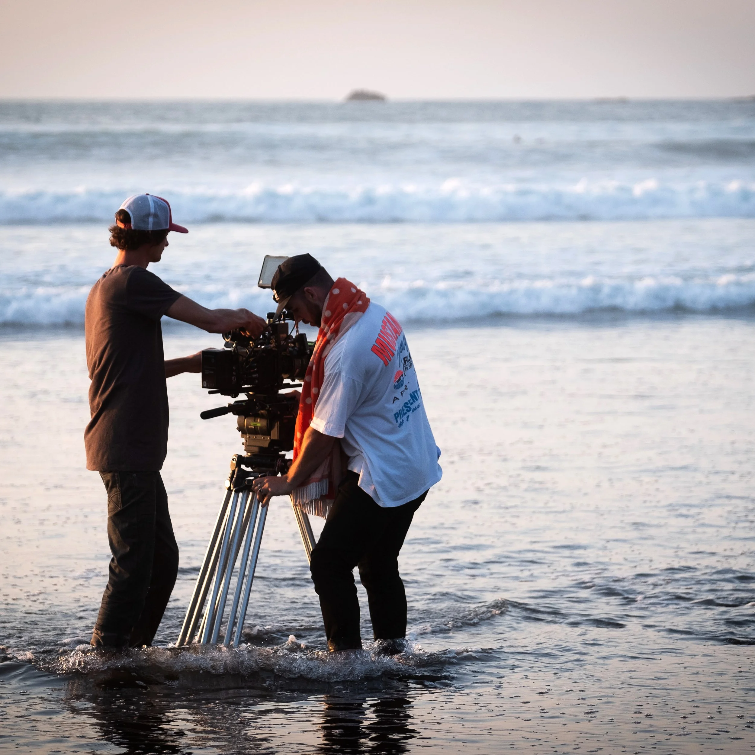 Two people filming with a professional camera on a beach at sunset, with the ocean and waves in the background.