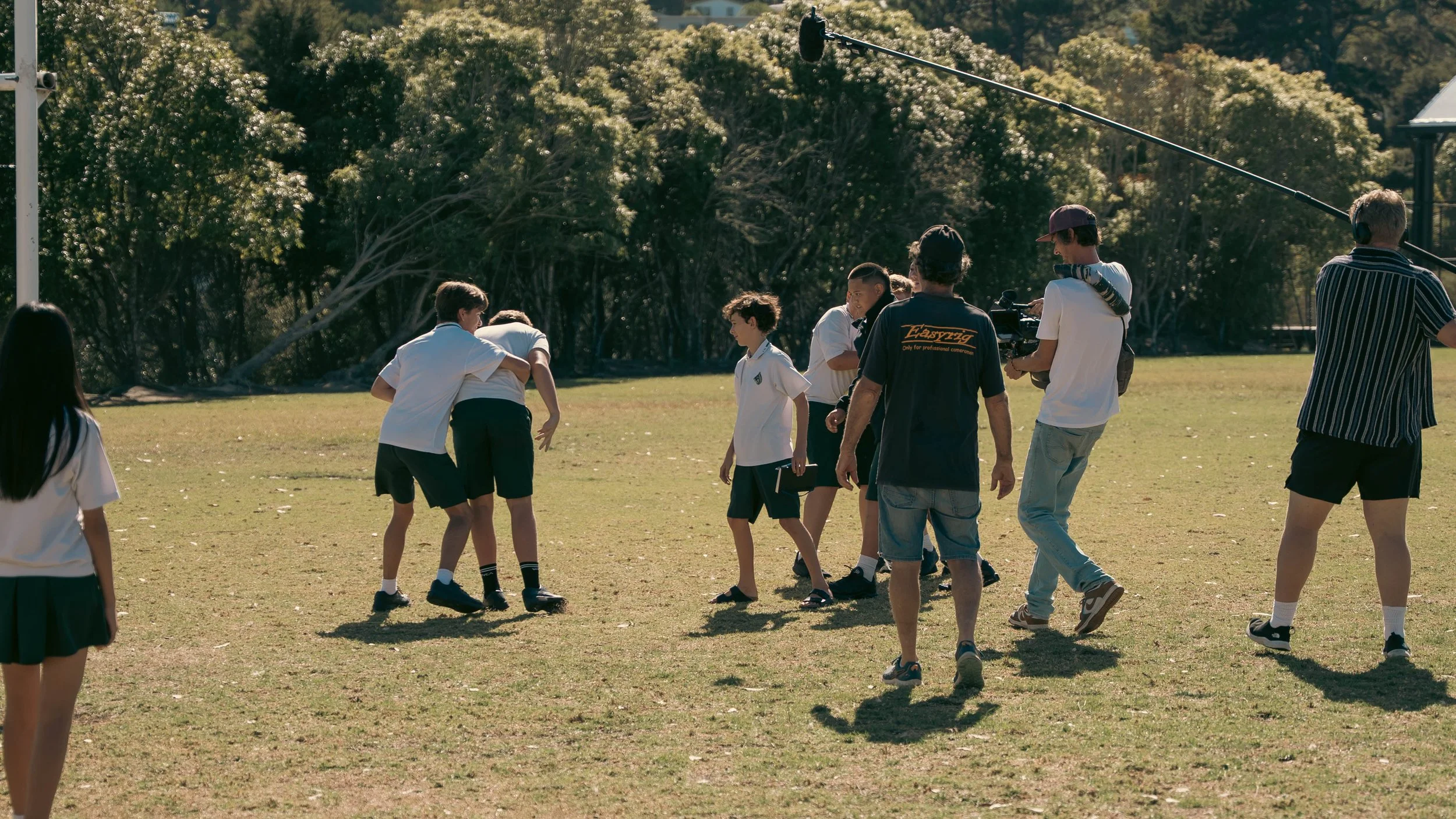 Children in school uniforms are on a field, with some being filmed by a crew, and a few are hugging or standing.