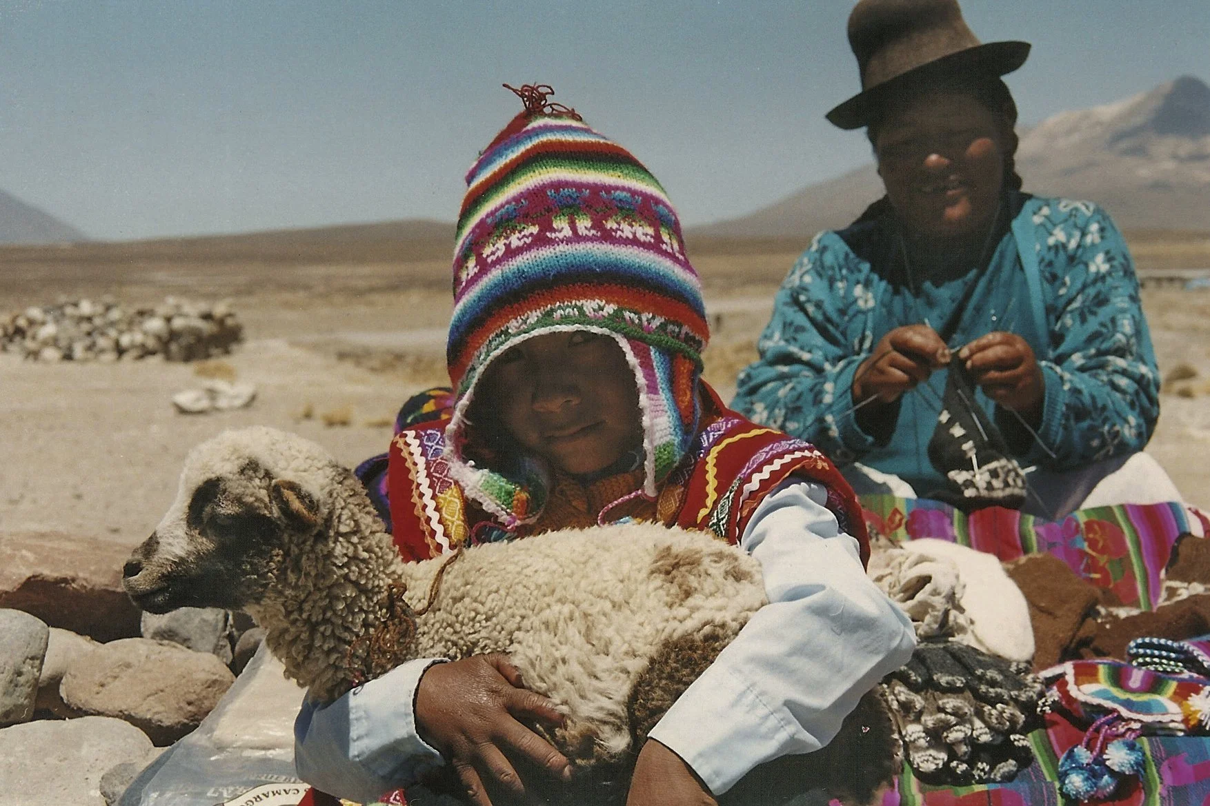 Two indigenous women wearing colorful traditional clothing and hats sitting on rocky ground in a desert landscape with mountains in the background, holding sheep.