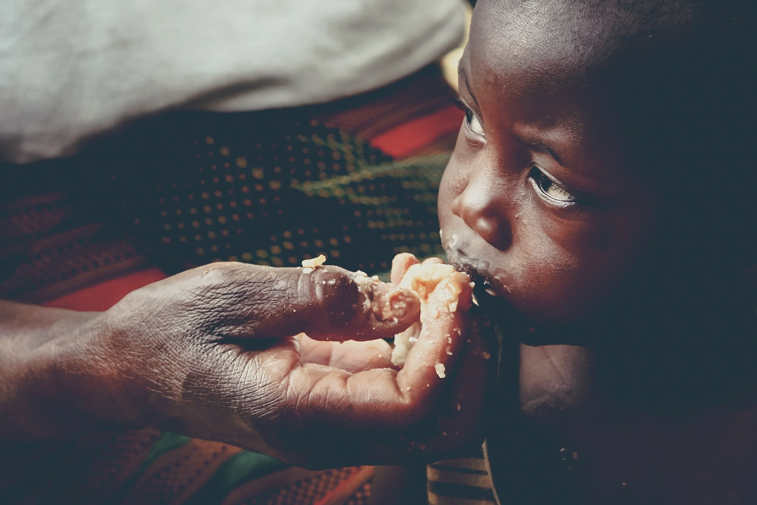 A young boy with dark skin is being fed food by an adult hand. The boy is gently biting into a piece of food held near his mouth.