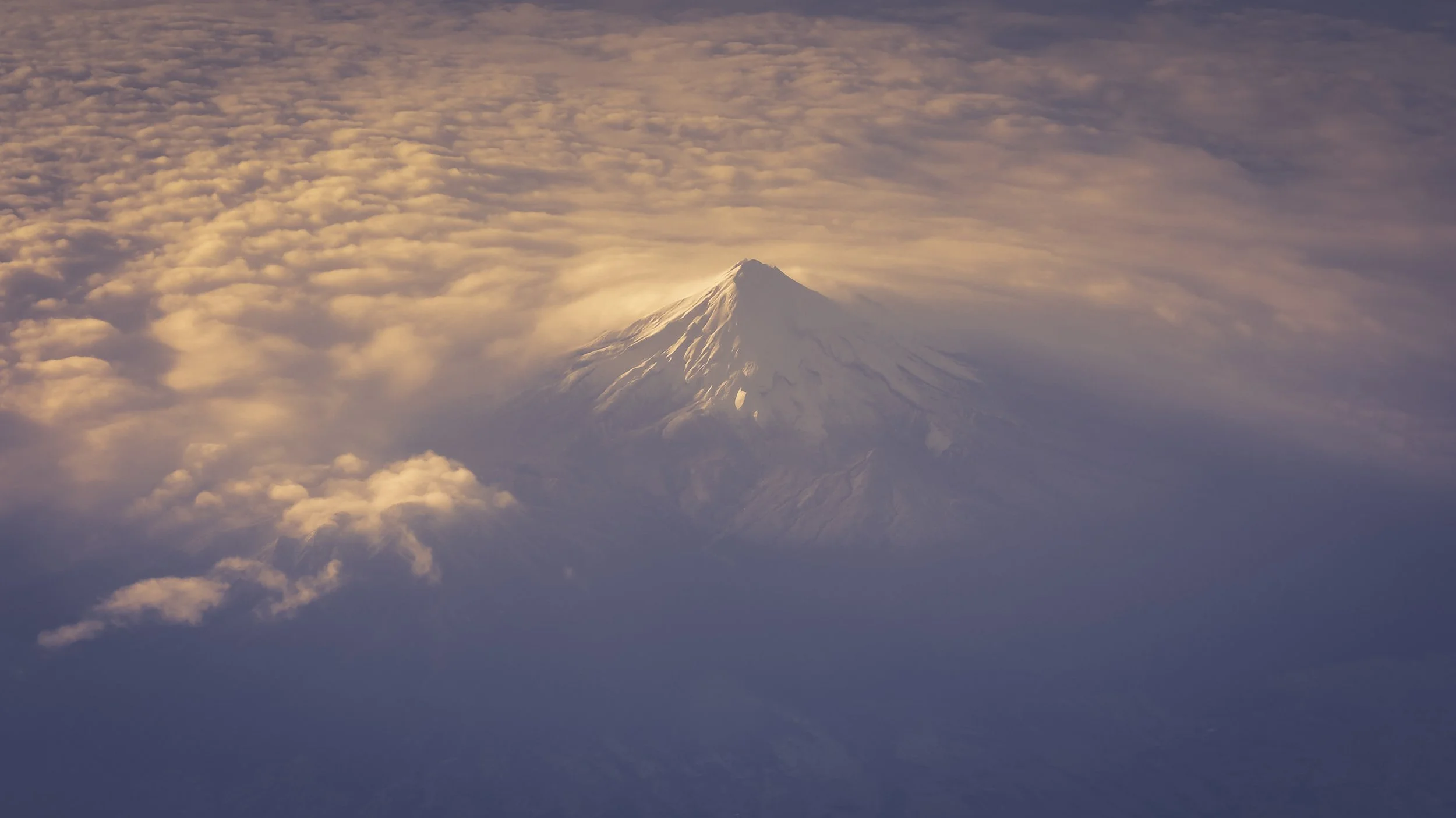 A snow-capped volcano rising above clouds during sunset or sunrise.