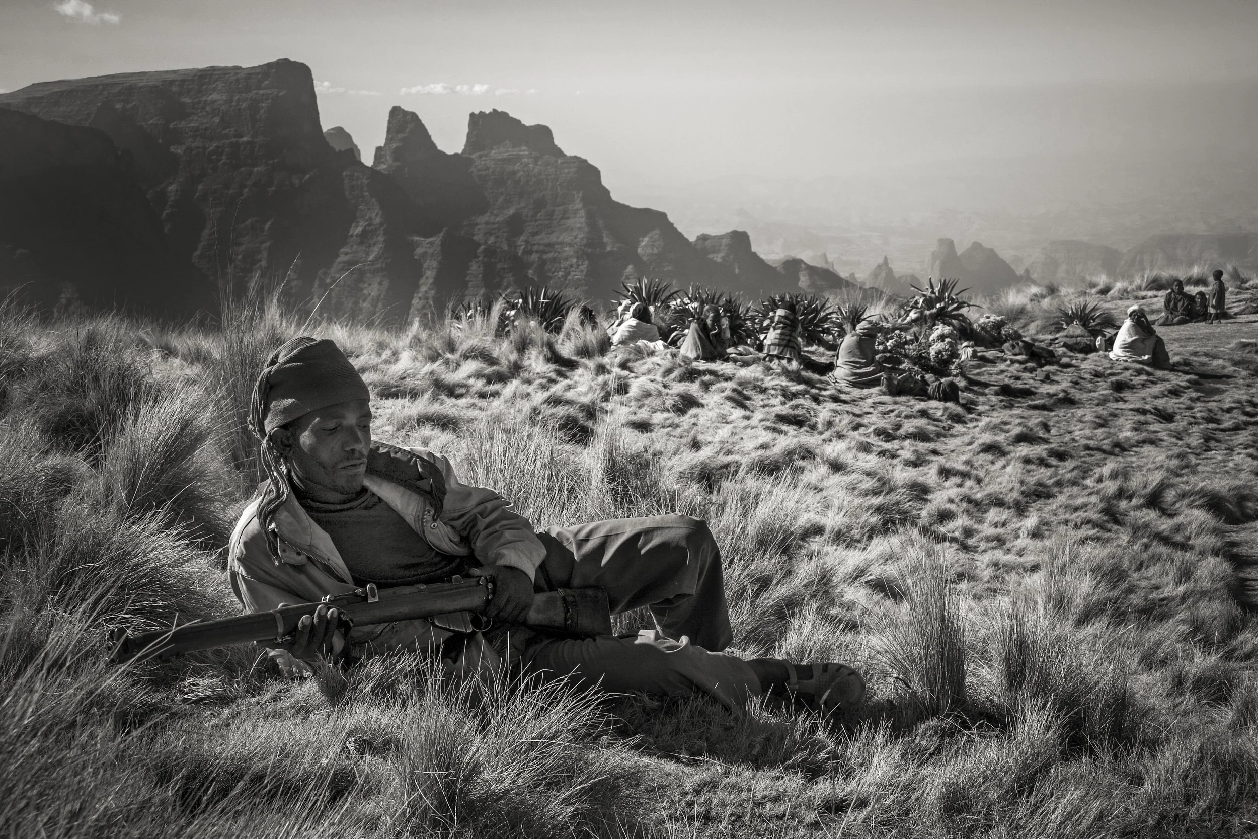 A man resting on the grass with a rifle, surrounded by a group of people sitting and standing among plants in a mountainous landscape.