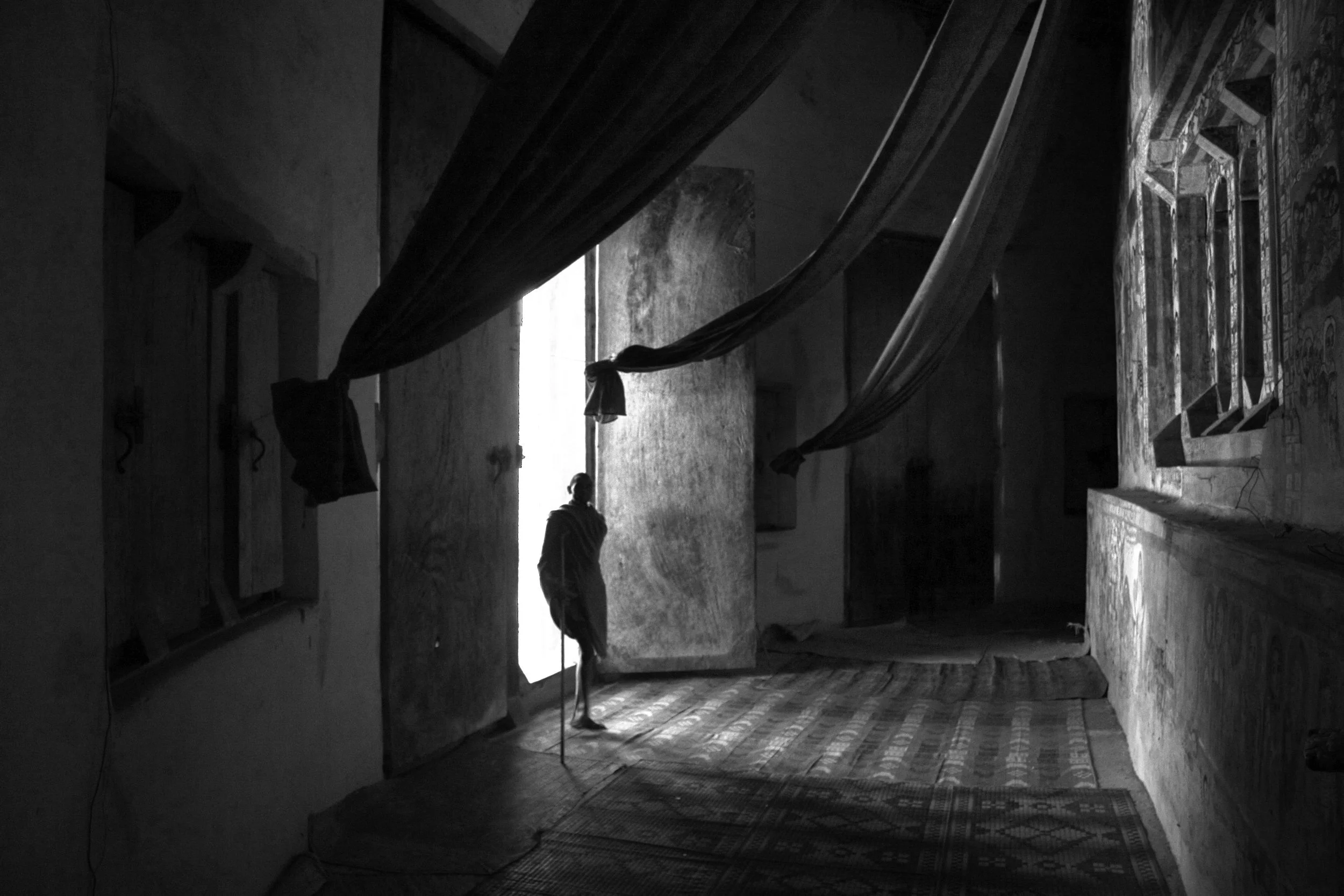 A silhouette of a person standing inside a dimly lit room, with light coming through an open door or window behind them. The room contains hanging fabric and patterned rugs on the floor, with textured walls and a painting or mural on the right wall.
