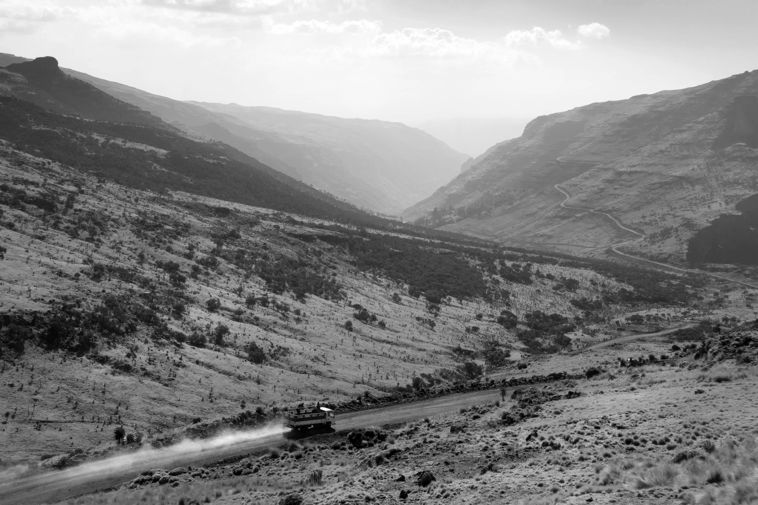 A black and white photo of a mountainous landscape with a dirt road running through the valley, and a bus traveling on the road kicking up dust.