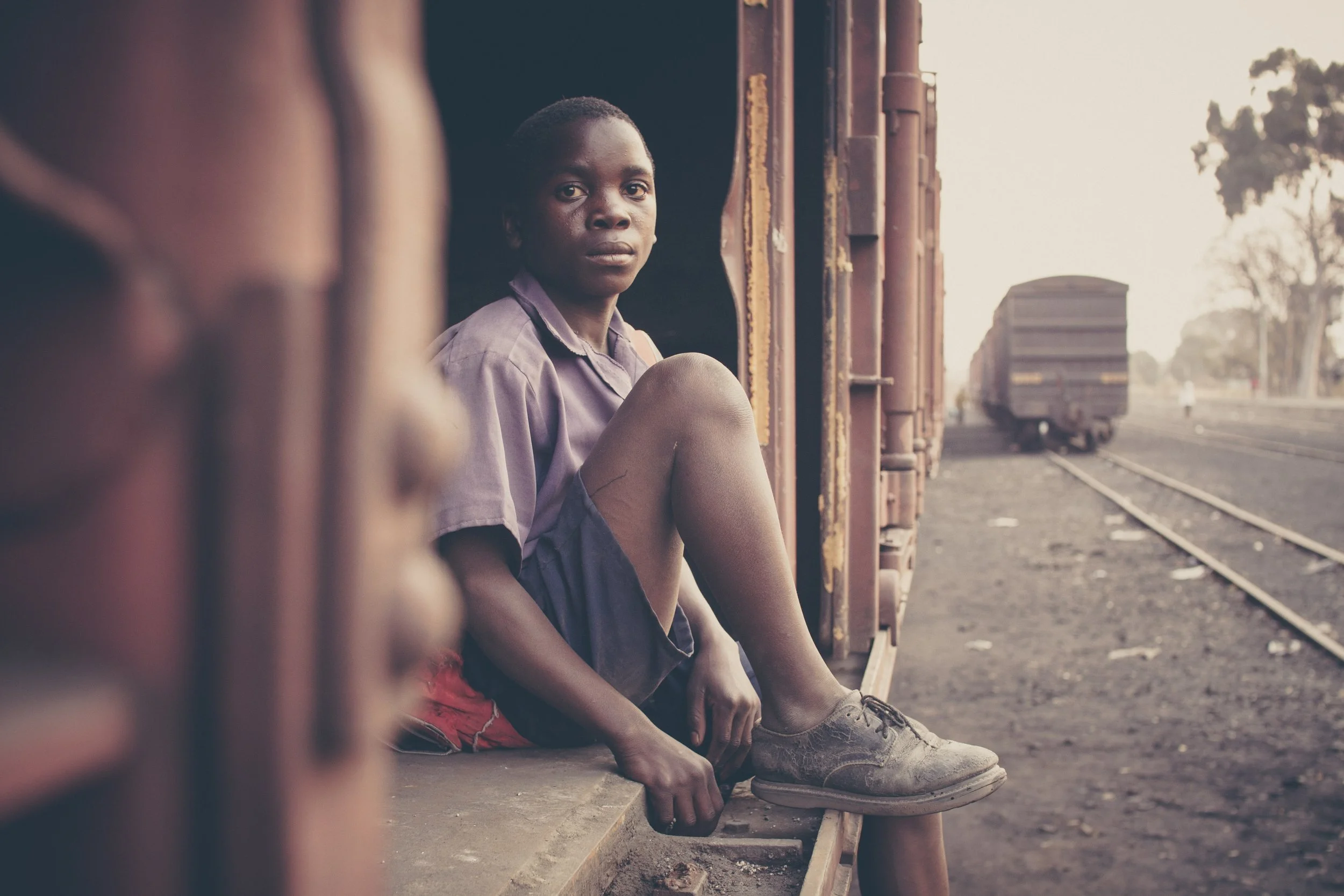 A young boy in a purple shirt and red shorts sitting on the edge of a train car, looking towards the camera. Train tracks and a blurred train are visible in the background.