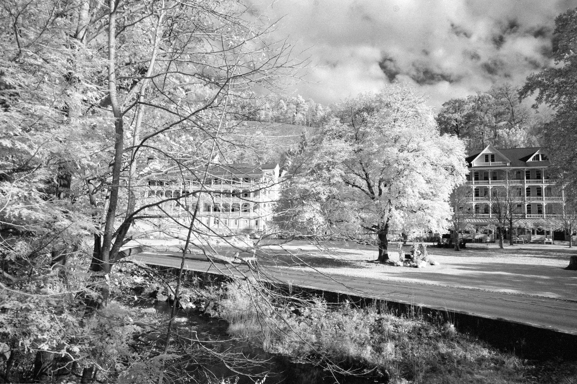 Black and white photo of a park with a small creek, leafless trees, and large houses with multiple balconies in the background under partly cloudy skies.