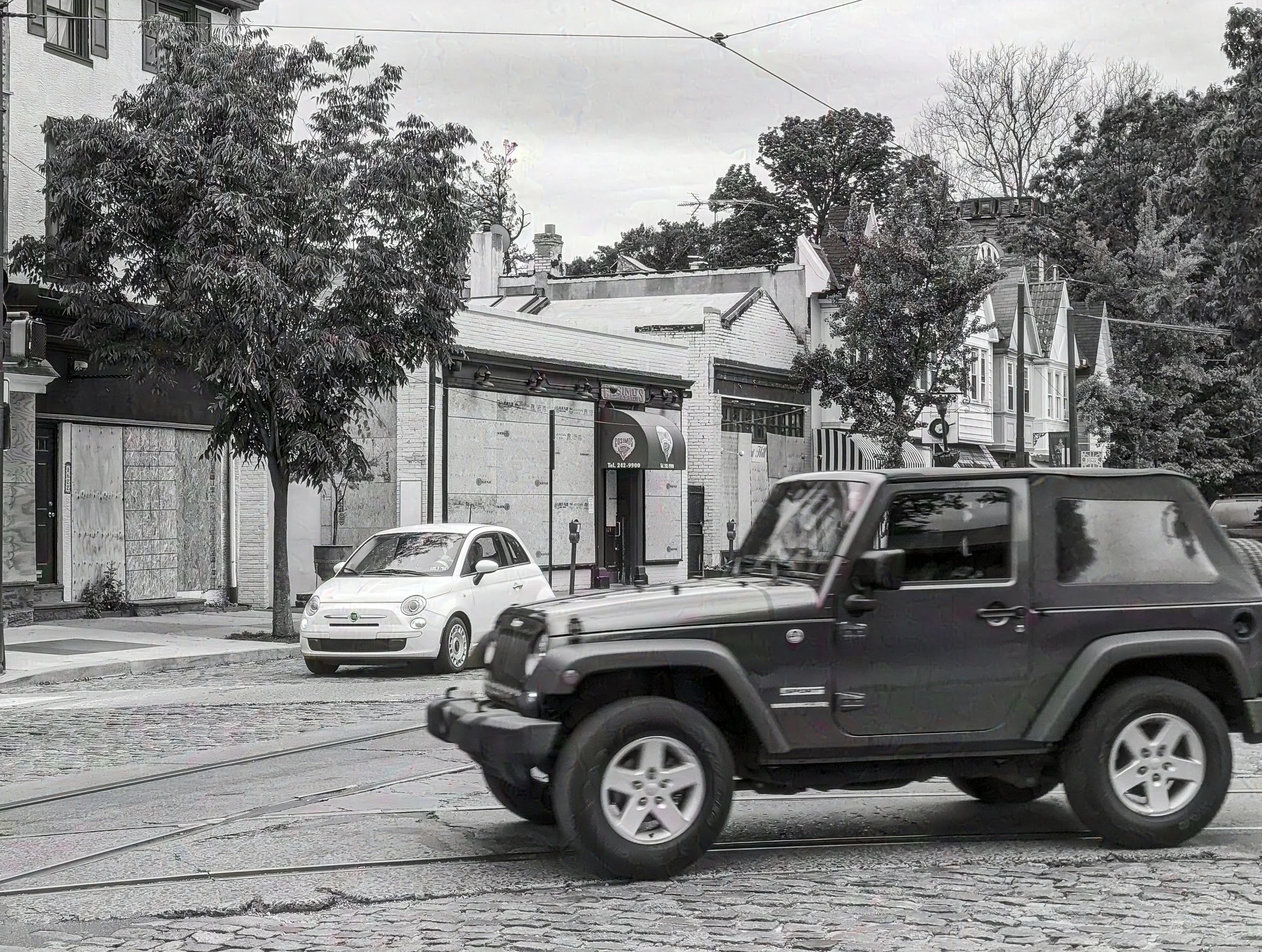 Black Jeep with a convertible roof driving on a brick street, with a small white car parked on the sidewalk and trees and buildings in the background.