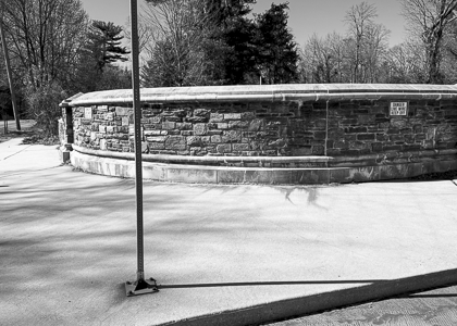 Stone wall alongside a paved driveway with a pole casting a shadow.