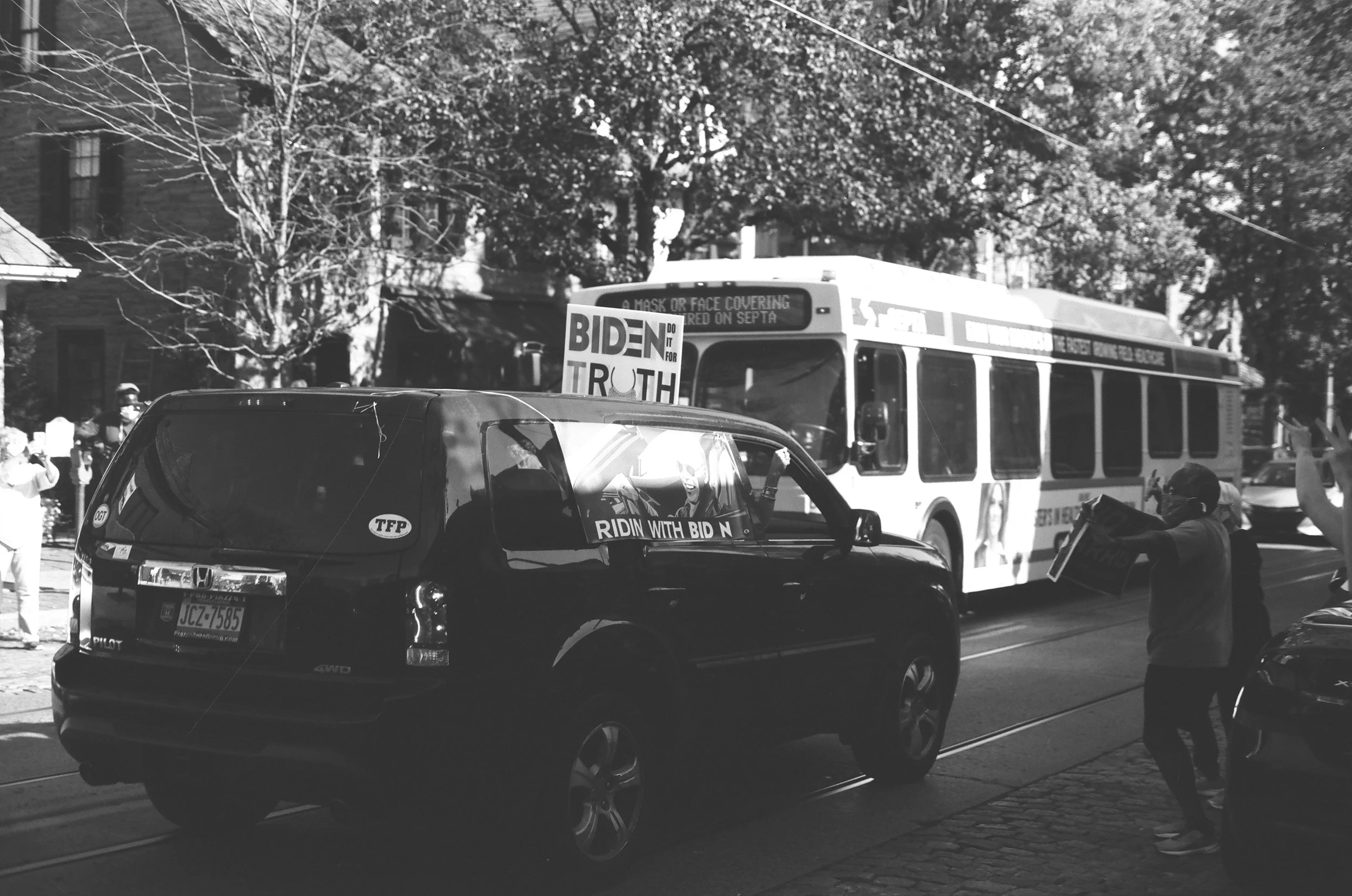 A street scene with protestors holding signs that read "Biden Do It For Truth" and "Ridn with Bid N." A bus displaying a message about face covering regulations and a person holding a sign. Several cars are visible, and there are trees and buildings 