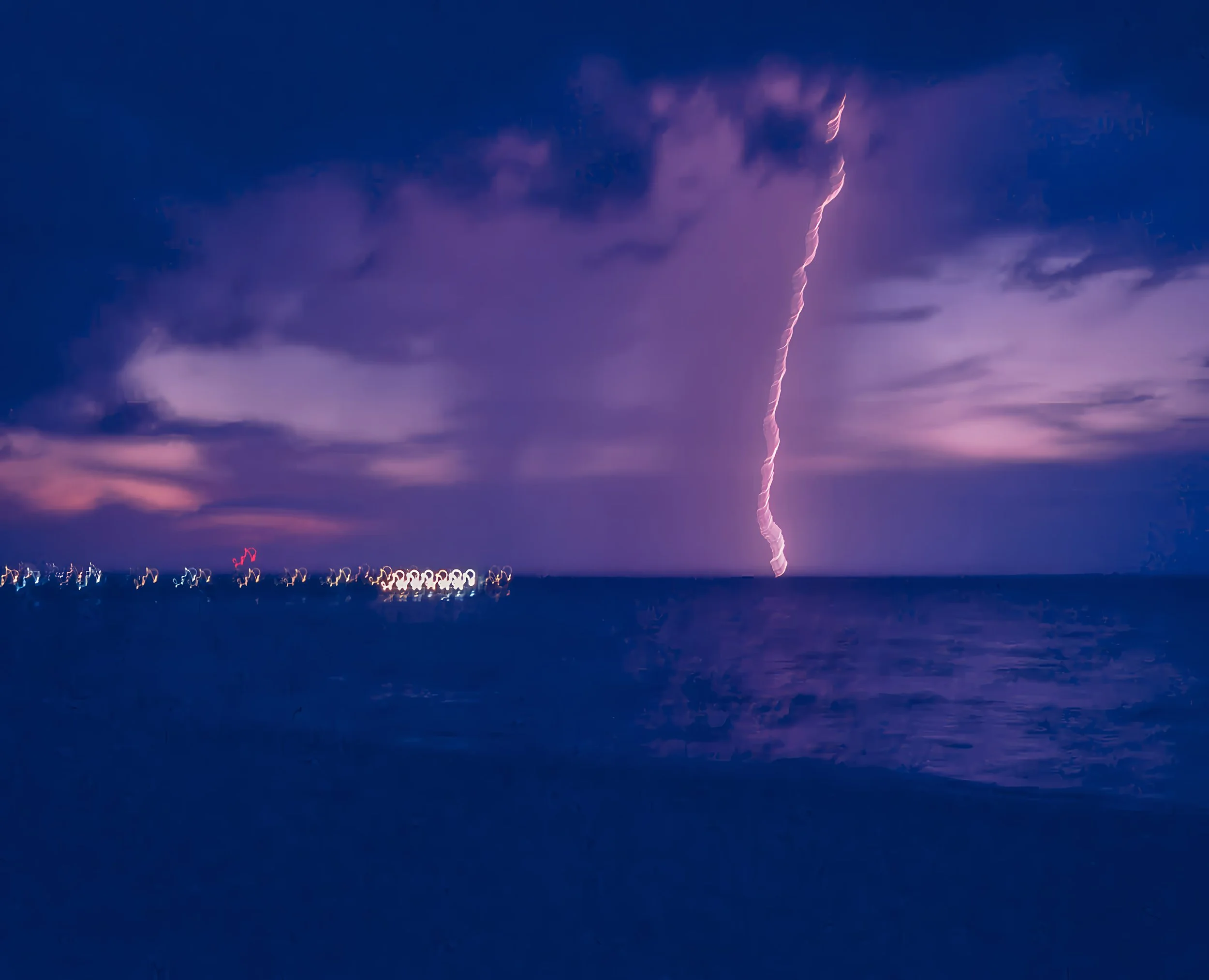 A lightning strike illuminating the night sky over the ocean with distant city lights on the horizon.