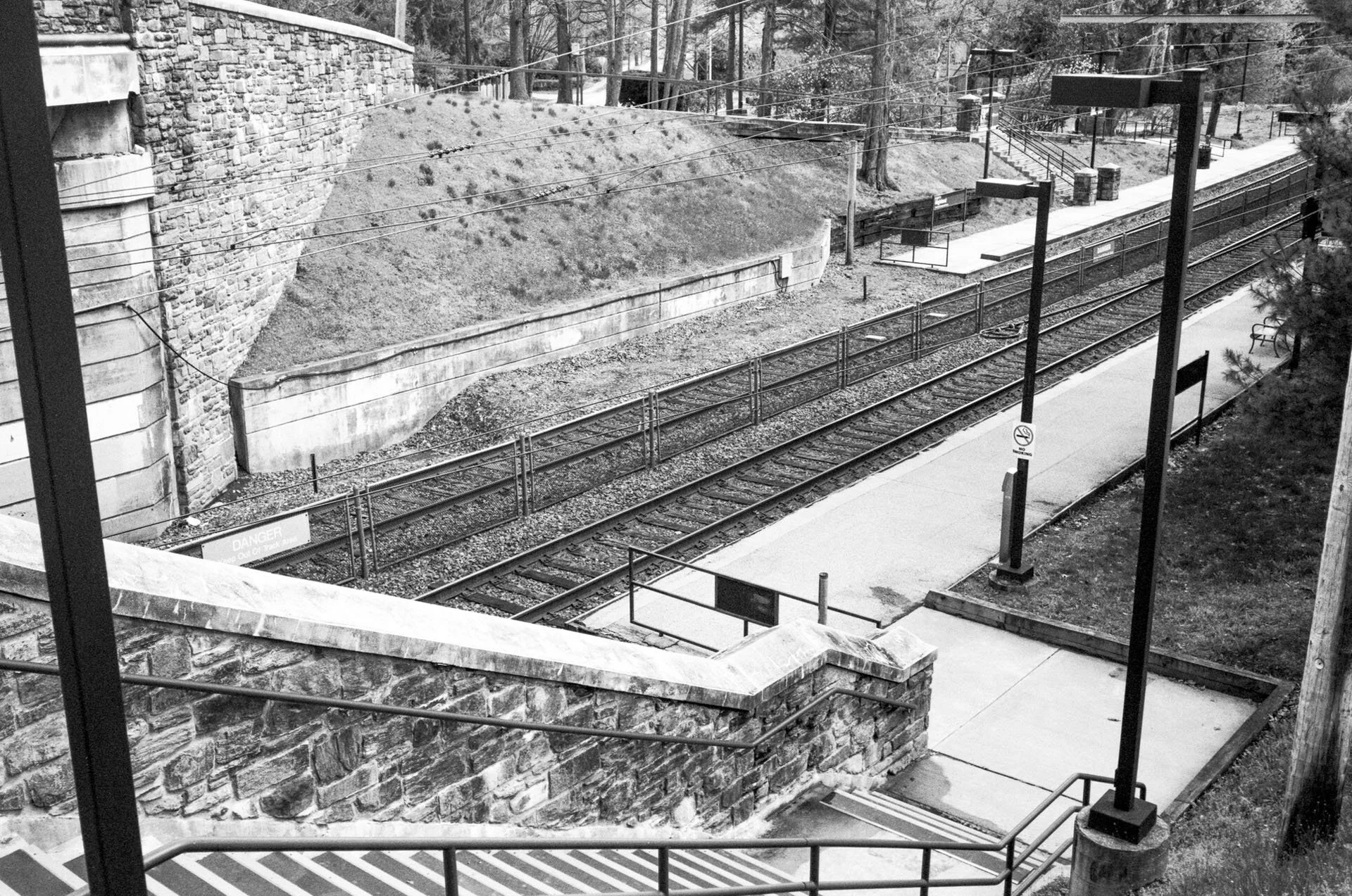 Empty outdoor train platform with multiple train tracks, stone and concrete walls, and tall lamp posts, black and white photo.