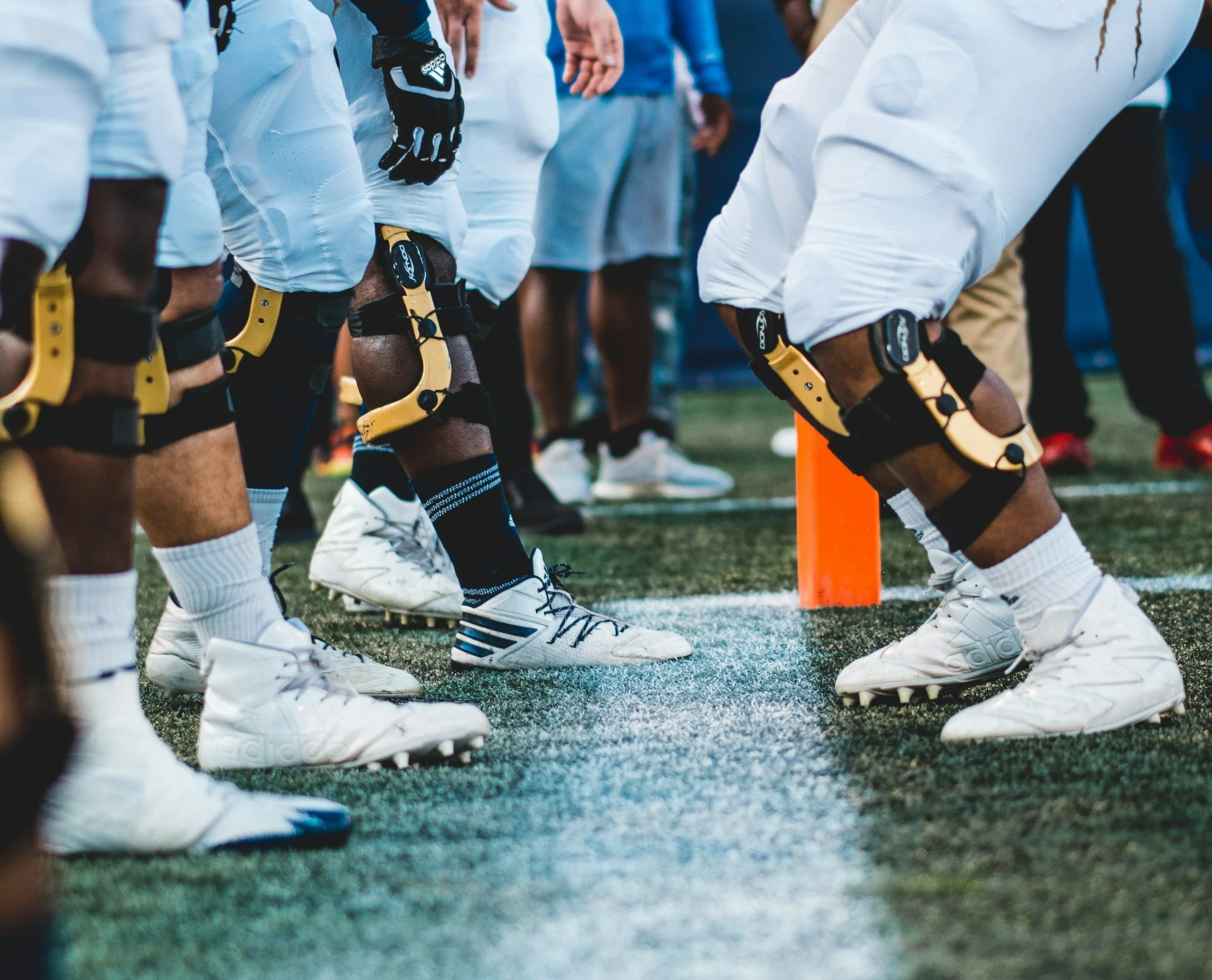 Flag football players in white uniforms wearing yellow knee braces, lined up on the field during a game with some players wearing black gloves and cleats.