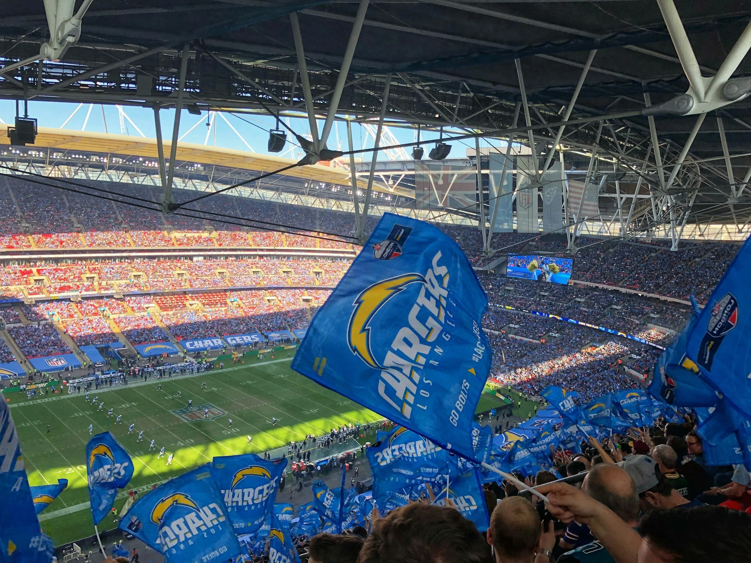 A football stadium filled with spectators, with fans waving blue Chargers flags, as a game is in progress on the field.