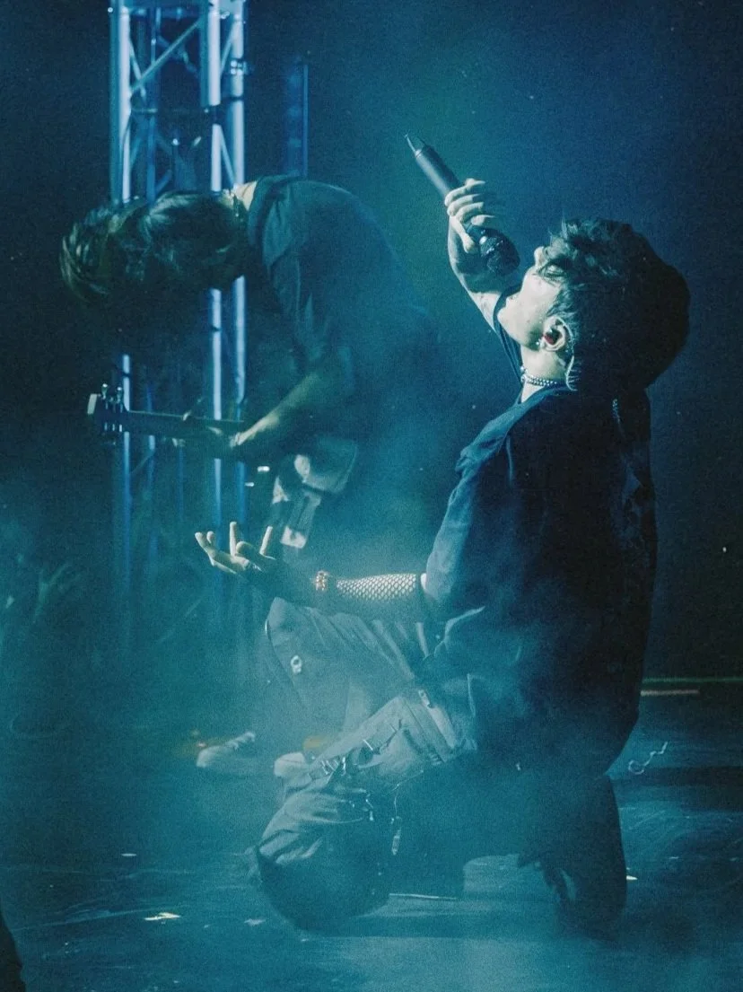 Musicians performing on stage with blue lighting and smoke, one person kneeling and singing into a microphone, another playing guitar, and a third with long hair leaning forward.